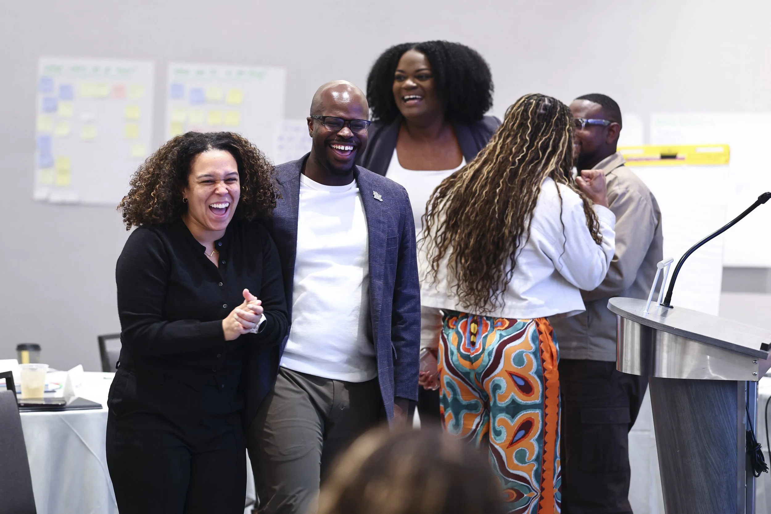 Group of five diverse people laughing and smiling at a professional event or conference, with a whiteboard and presentation materials in the background.