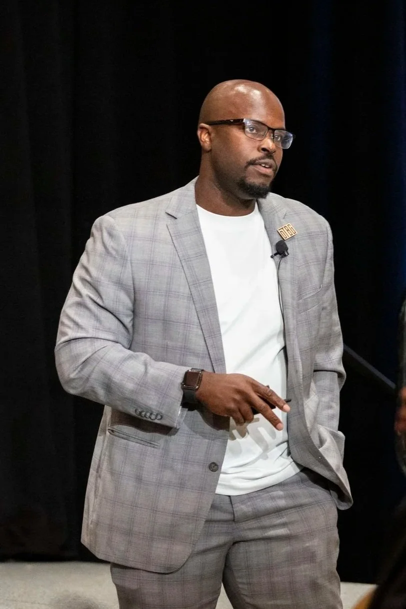 A man wearing glasses and a gray plaid suit jacket with a white T-shirt underneath, standing against a dark background. He has a wristwatch and a lapel pin, and is pointing with his right hand.