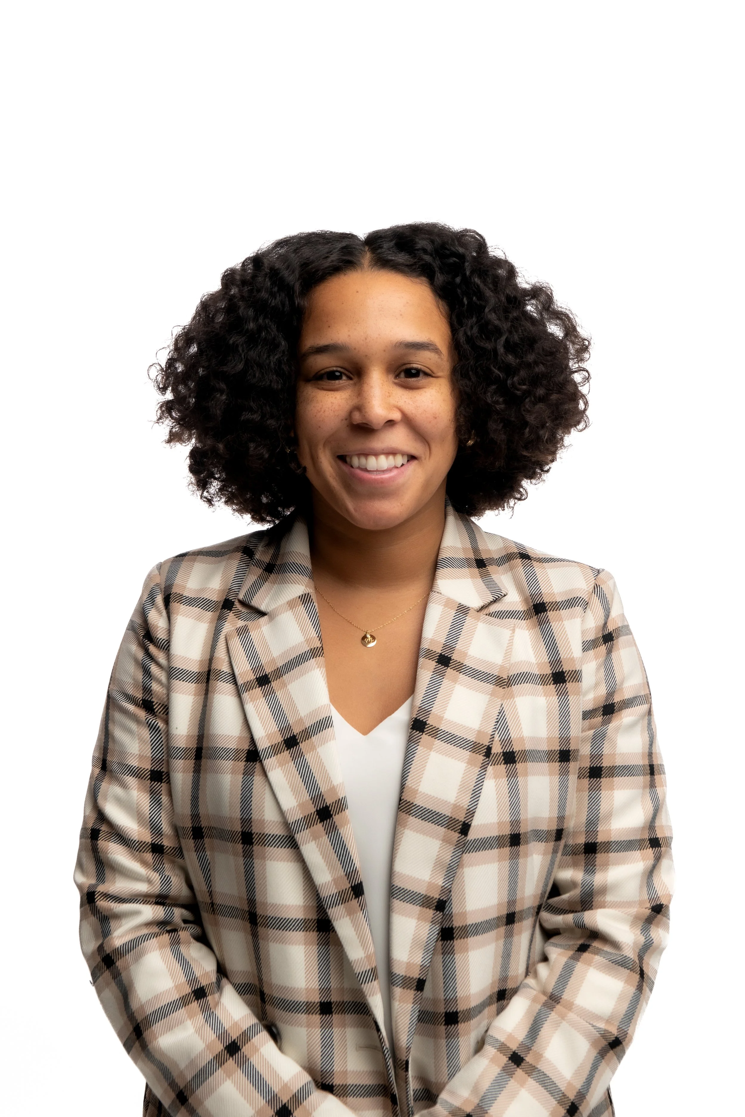 A woman with curly black hair smiling, wearing a beige and brown plaid blazer over a white top, against a plain white background.