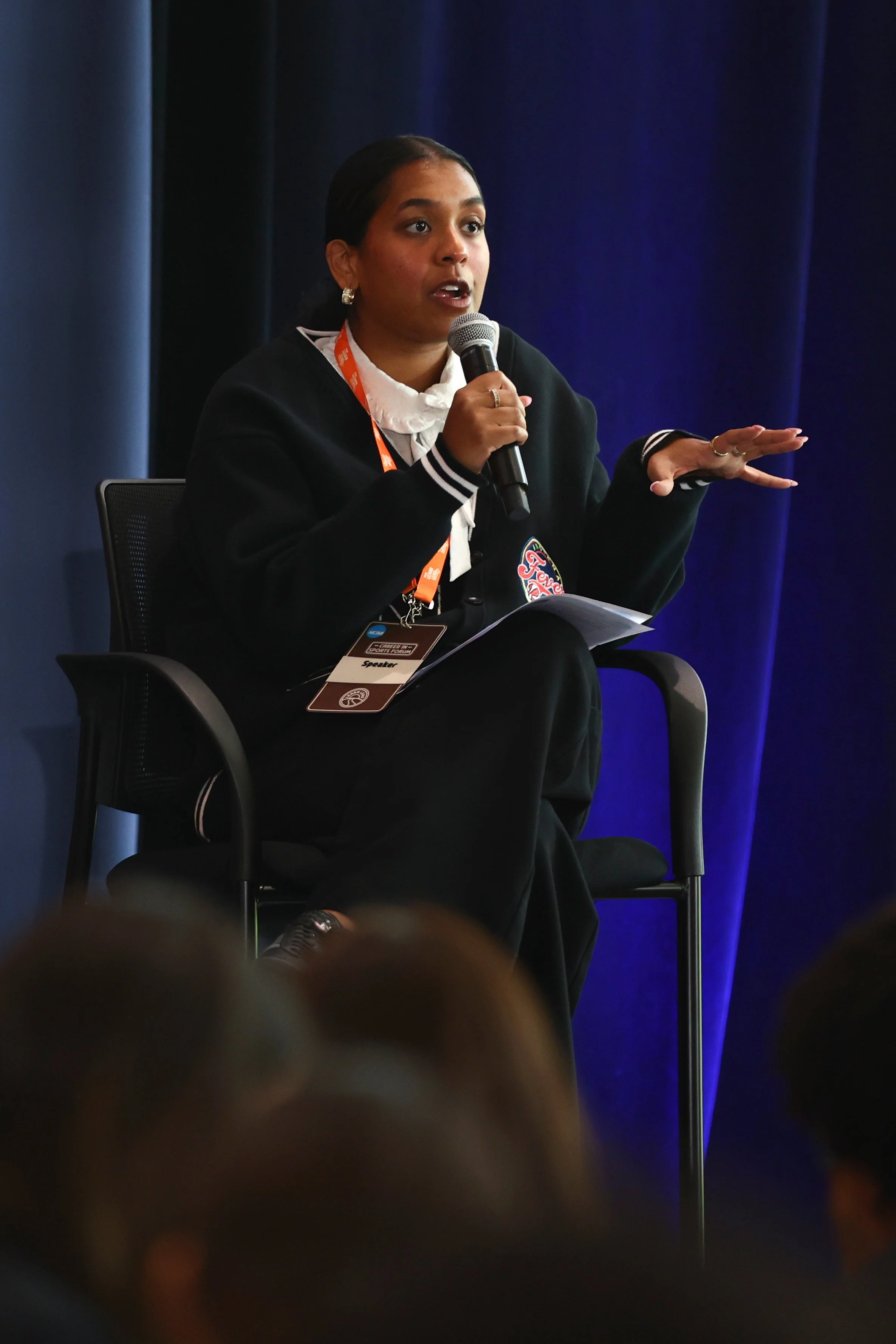 A woman speaking into a microphone during a panel discussion or conference. She is seated in a black chair, wearing a black jacket with a badge or logo, and a white shirt with a black ribbon. The background features dark blue curtains.