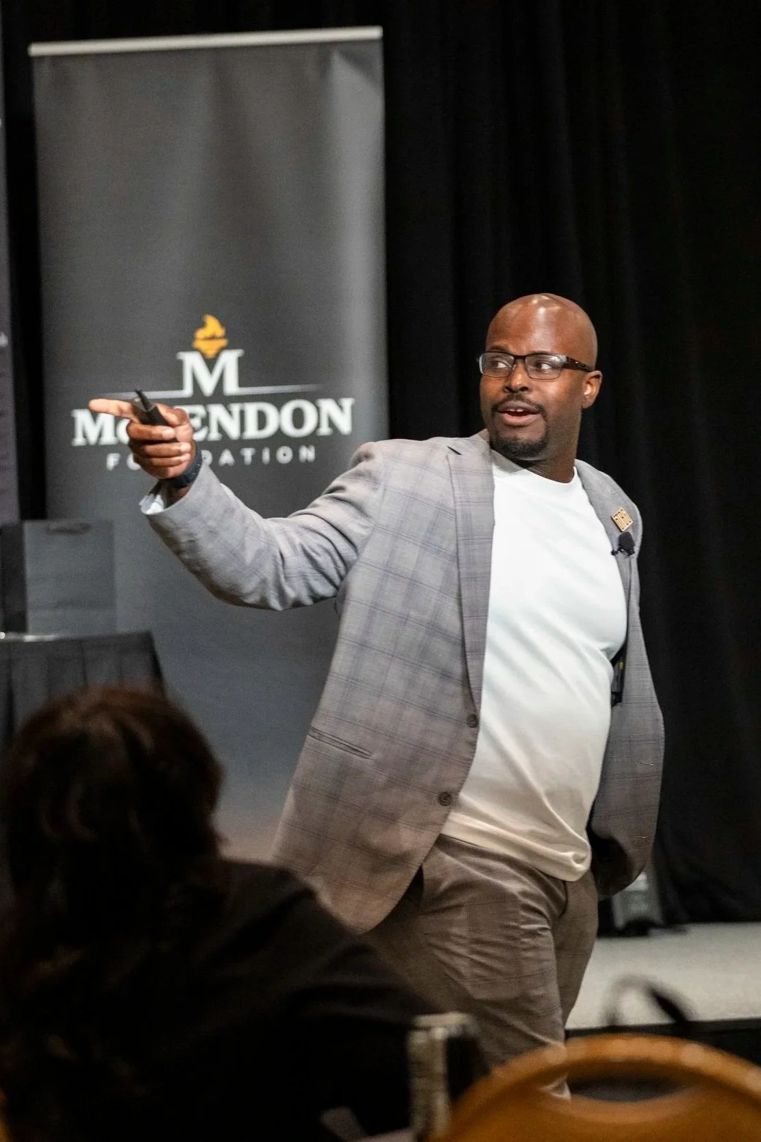 A man in glasses and a plaid blazer gestures while speaking at a presentation with a backdrop that reads "Mendon Foundation."