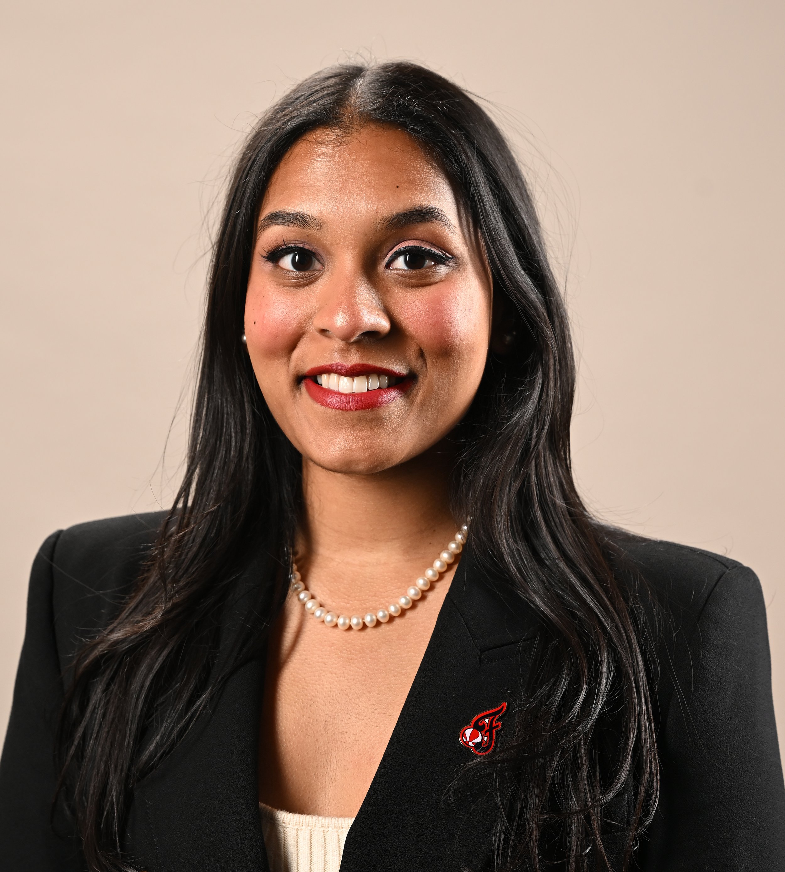Close-up of a woman with long dark hair, wearing a pearl necklace and a black blazer with a red logo, smiling against a plain beige wall.