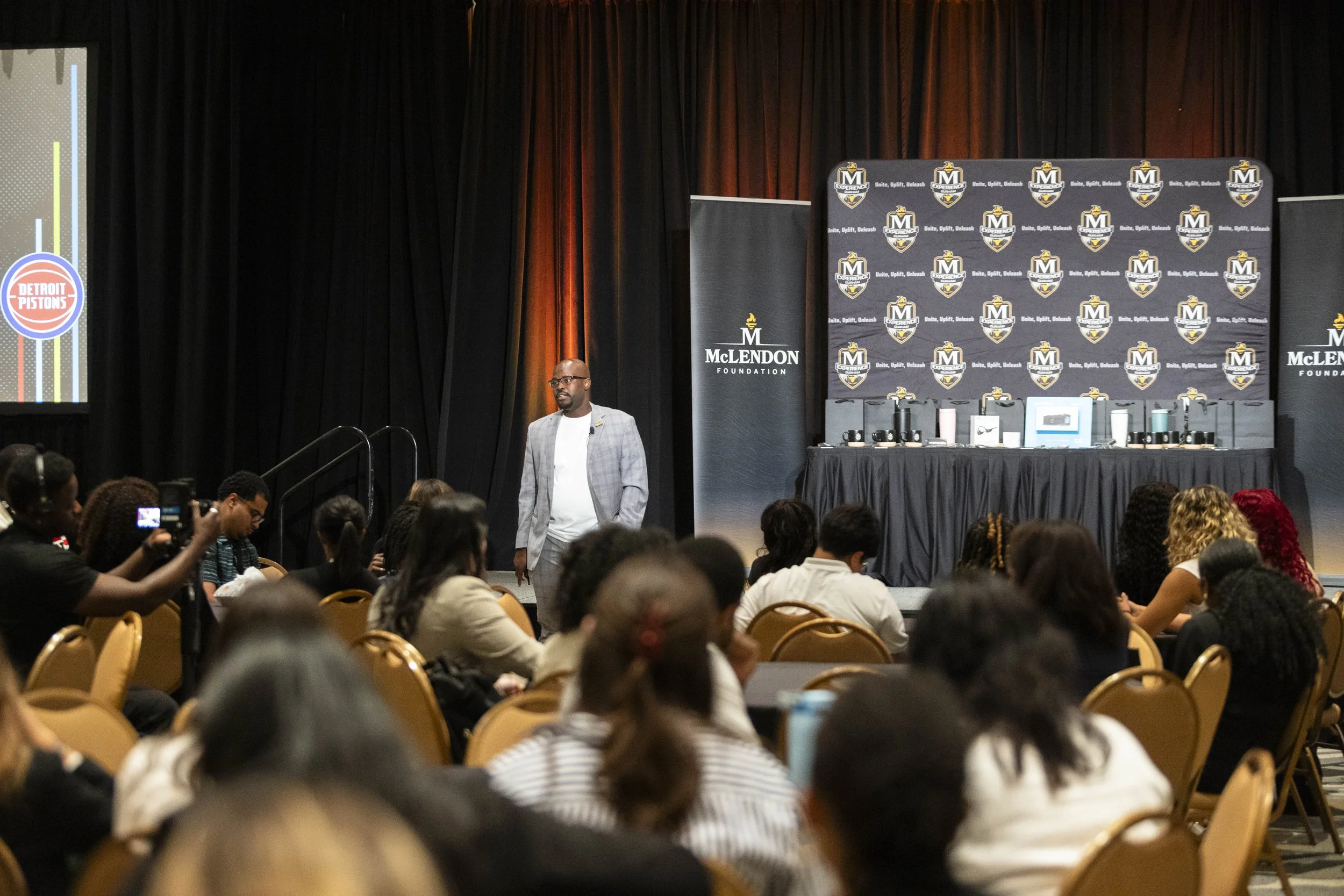 A man in a gray blazer and glasses is standing on a stage at an event with a McLendon Foundation backdrop. The audience is seated in rows of chairs, and there are two large screens displaying event logos and branding.