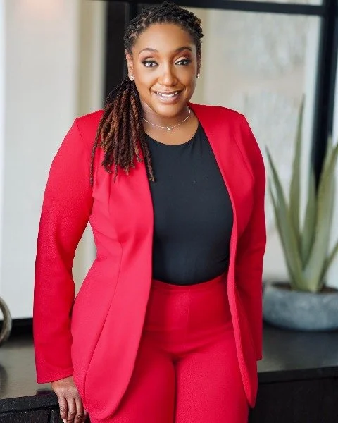 A confident woman with braided hair, wearing a red blazer and matching red pants, standing indoors near a window with a potted plant in the background.