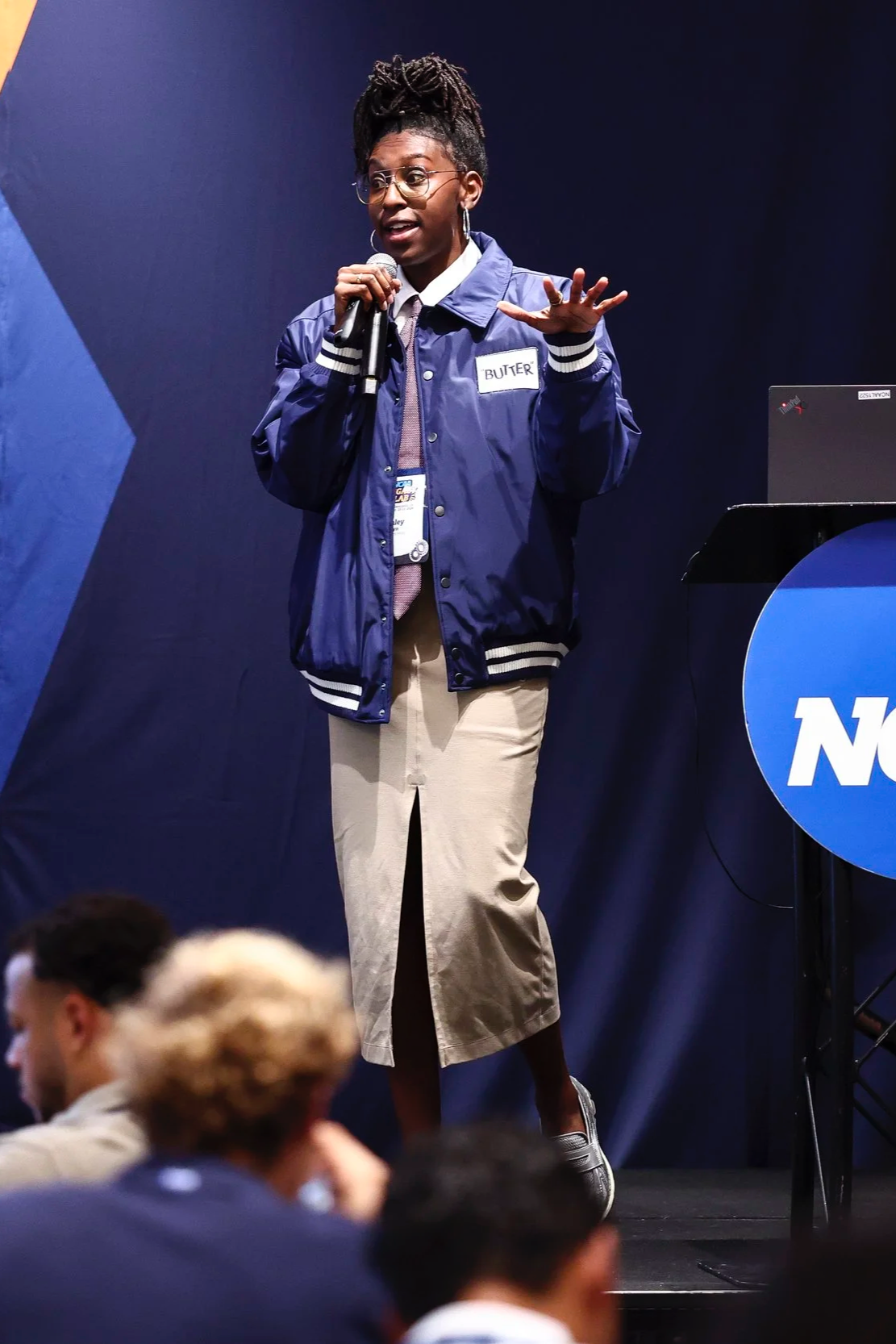 A woman with glasses and dreadlocks speaking into a microphone on stage at an event. She wears a blue jacket with a name tag that says 'Buter,' a white shirt, and beige pants. There is a laptop on a stand nearby.