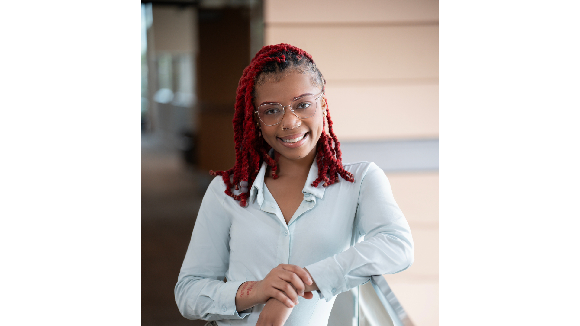 A young woman with red dreadlocks, glasses, and a septum piercing, smiling and standing with arms crossed in an indoor space.