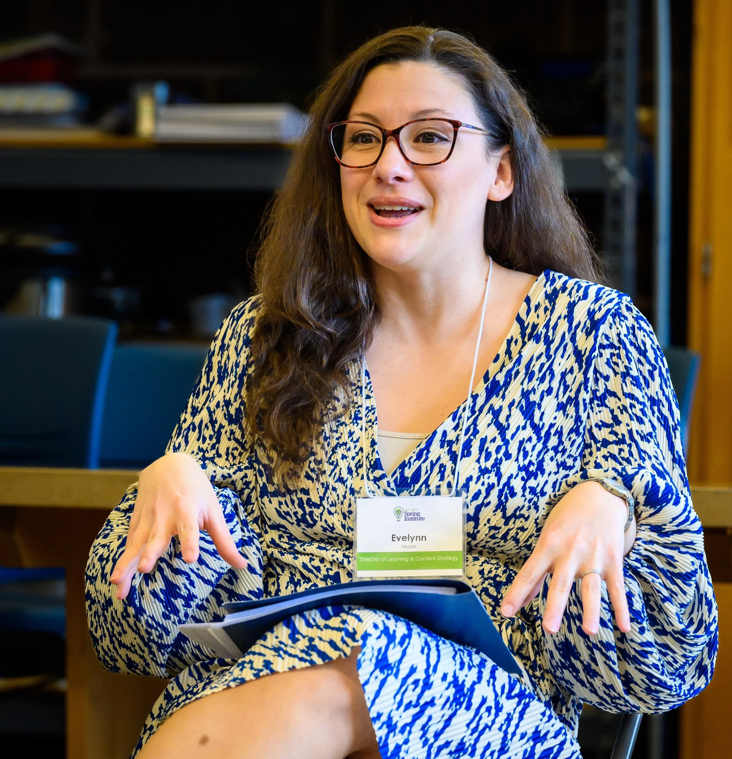 Woman with glasses, wearing a patterned dress, sitting and speaking at a conference or meeting, with a name tag that reads Evelynn Moon, Director of Learning & Content Strategy, at an indoor setting.