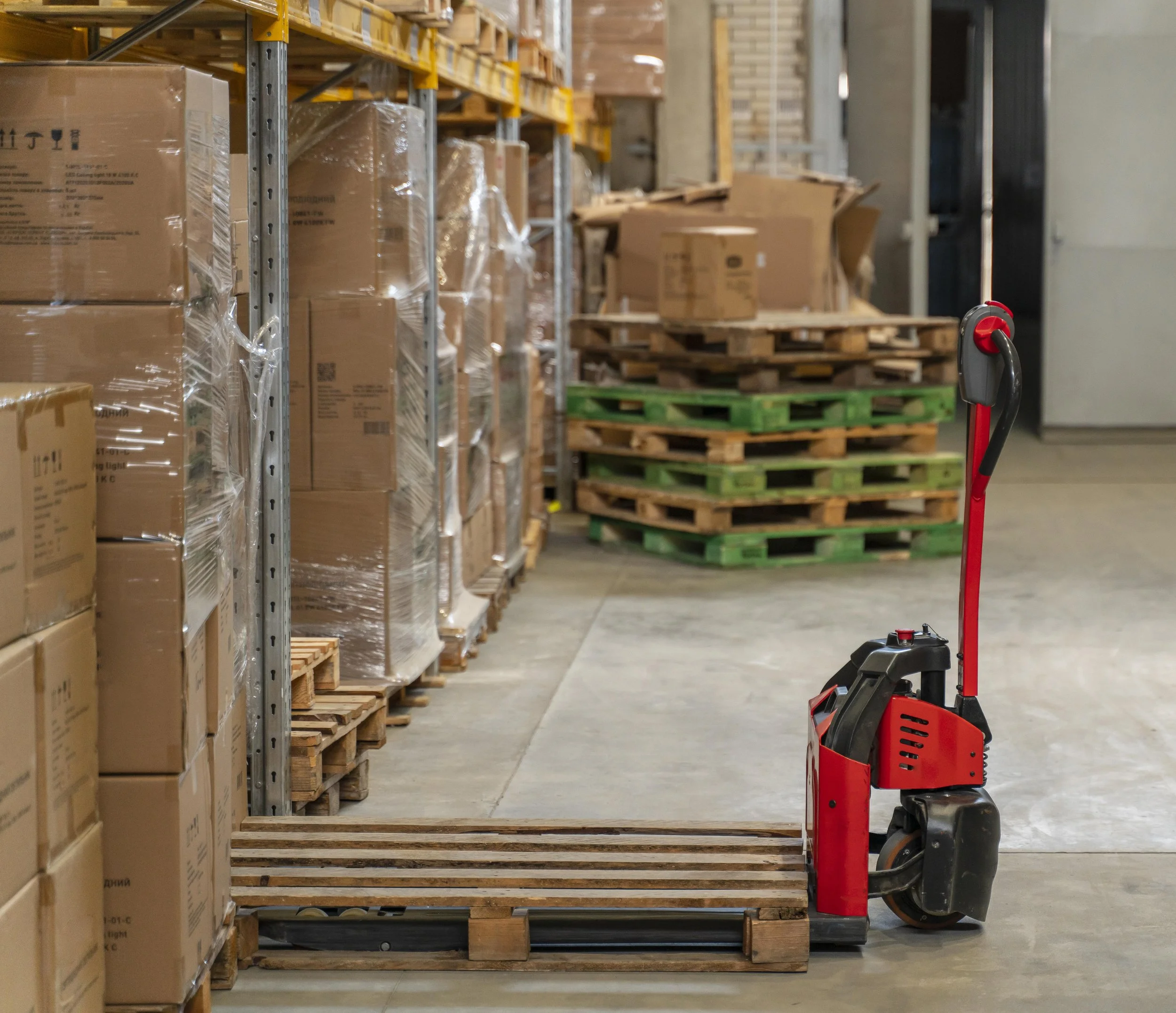 A red electric pallet jack in a warehouse, surrounded by stacked boxes on shelves and wooden pallets.