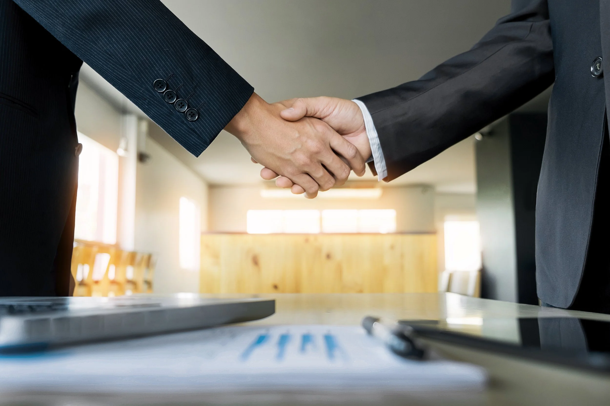 Businesspeople shaking hands in a meeting room with documents and a pen on the table.