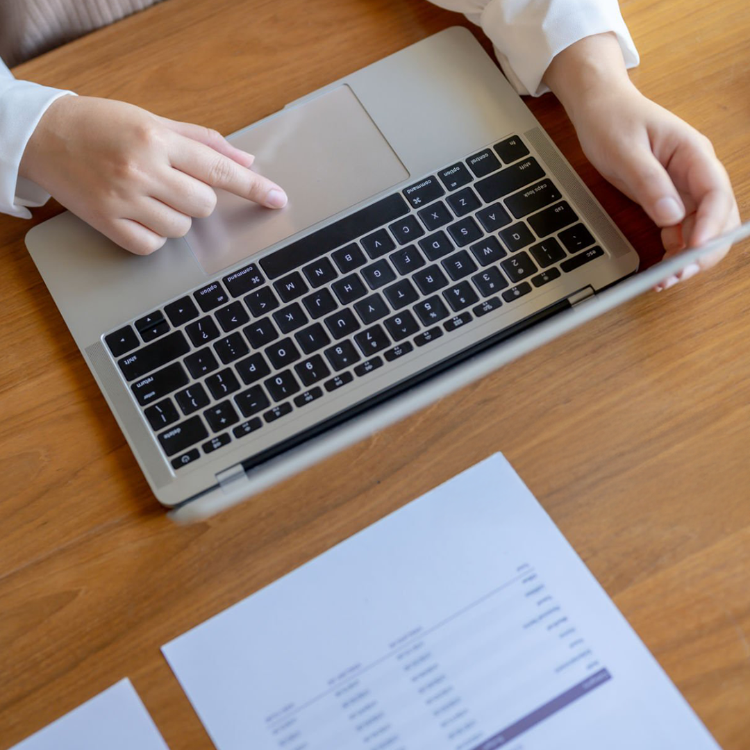 Person using a silver laptop on a wooden desk, pointing at the keyboard with one finger, with printed documents nearby.