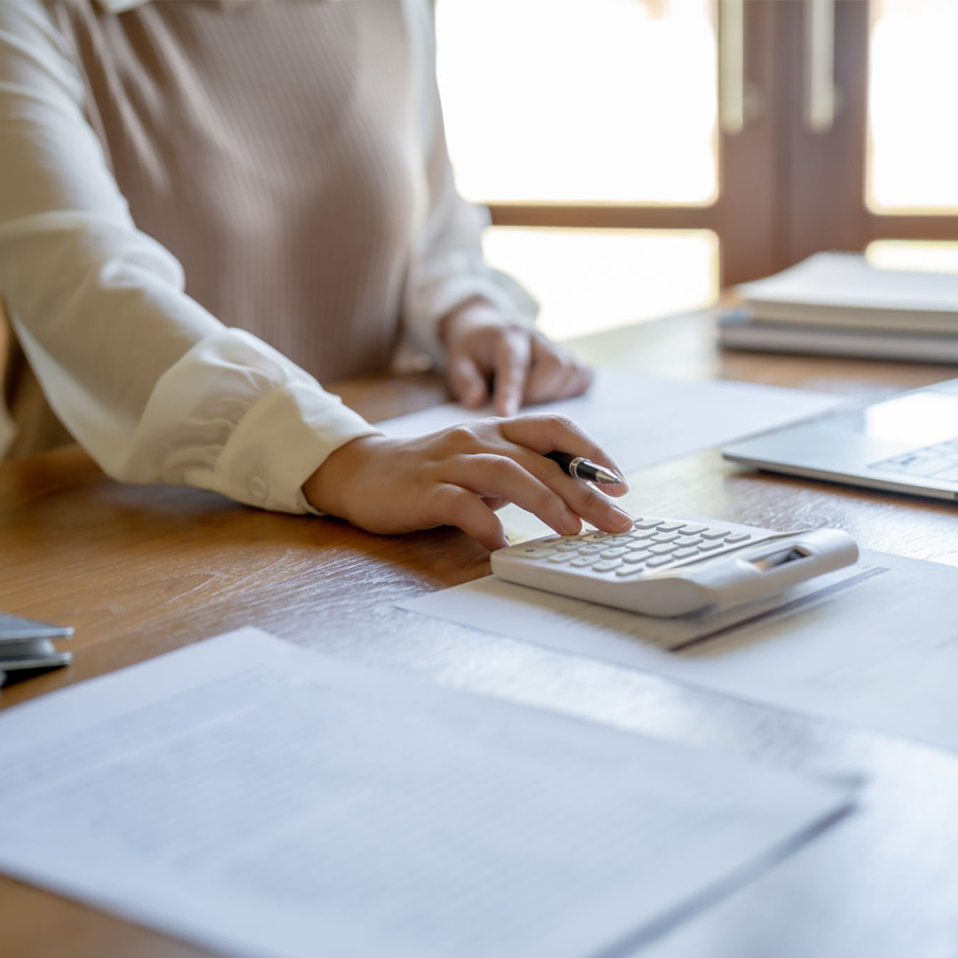Person using calculator on wooden desk with papers, pen, and laptop near window.