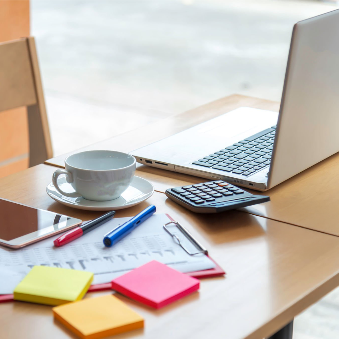 Work desk with a chair, a laptop, a white coffee cup on a saucer, a calculator, a tablet, multiple colorful sticky notes, and pens, on a wooden table near a window.