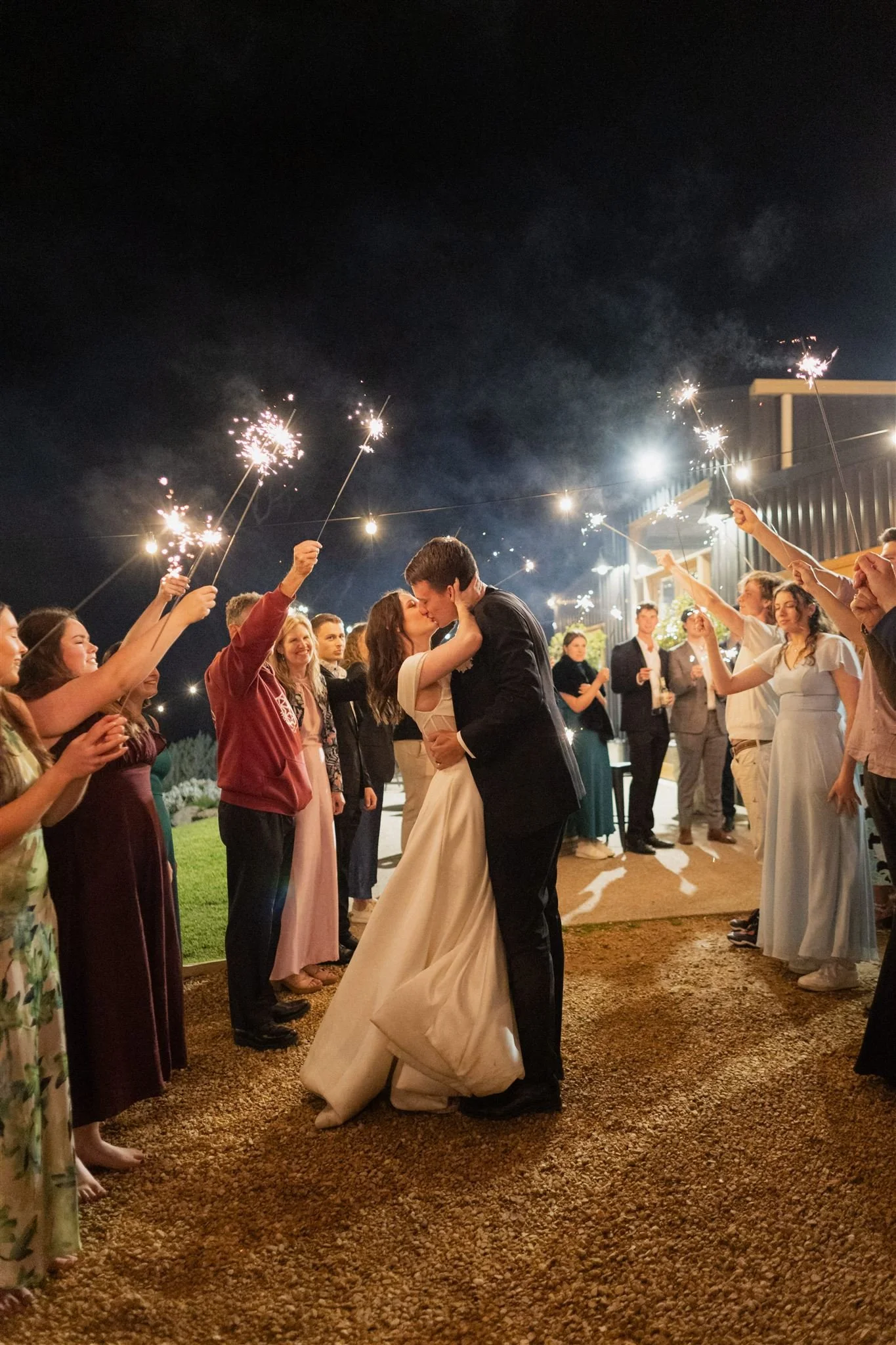 A newlywed couple sharing a kiss at night, surrounded by friends holding sparklers in the air during a celebration outside a building.