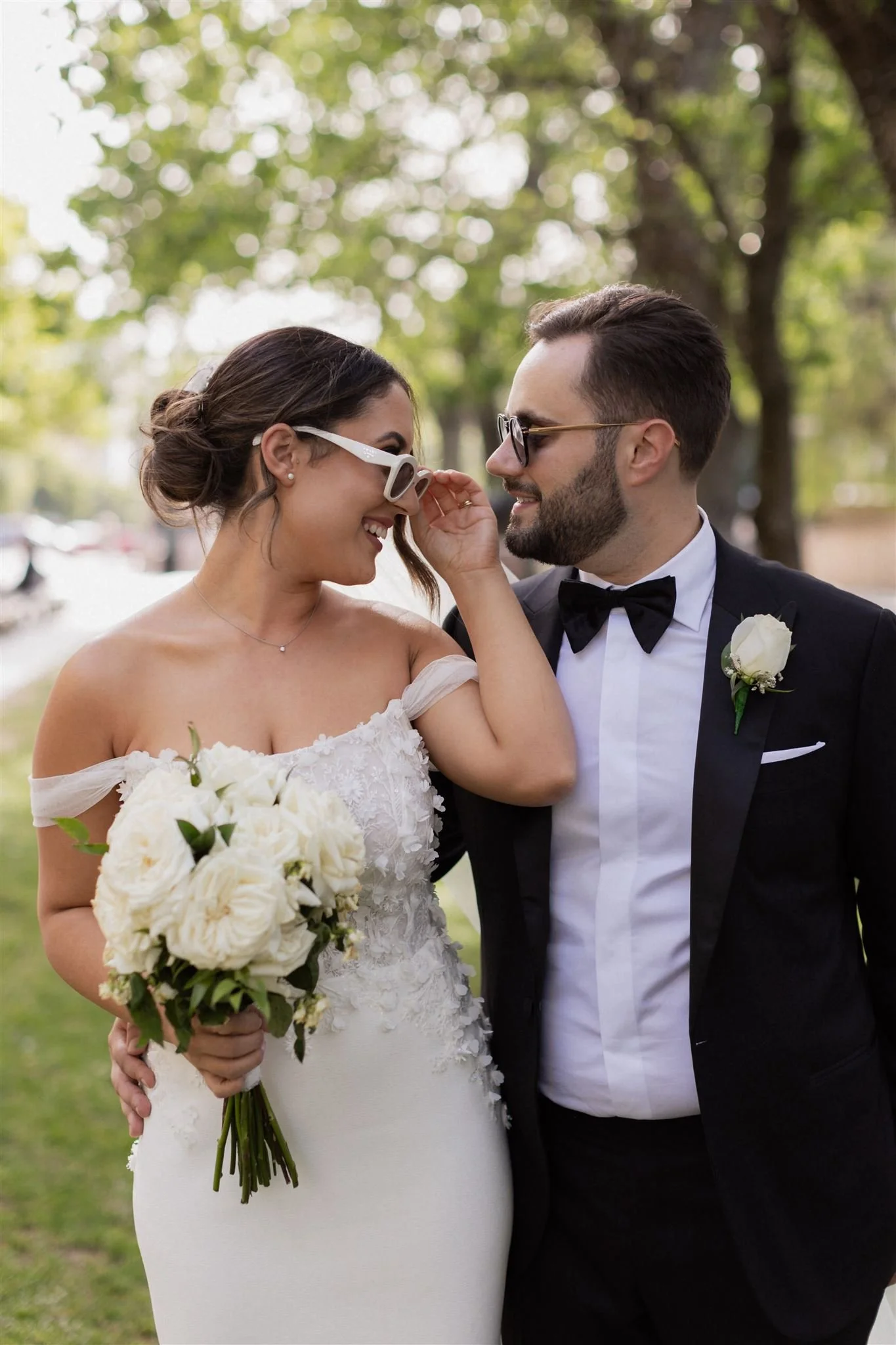 A bride and groom sharing a moment at their wedding outdoors, with the bride wearing sunglasses and holding a bouquet of white flowers, and the groom adjusting her sunglasses.