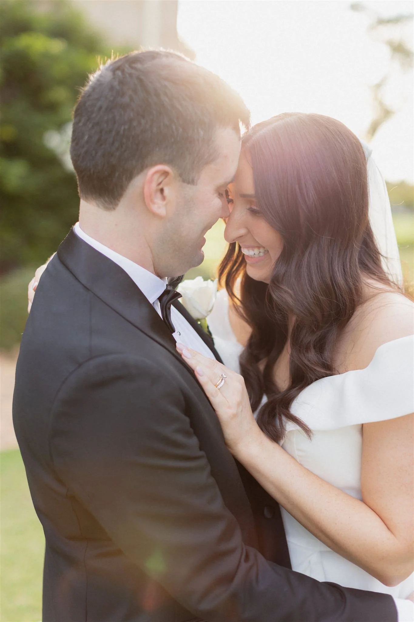 A newlywed couple sharing a joyful moment outdoors with their foreheads touching and smiling, the groom dressed in a black tuxedo and the bride in a white wedding dress and veil, with sunlight in the background.