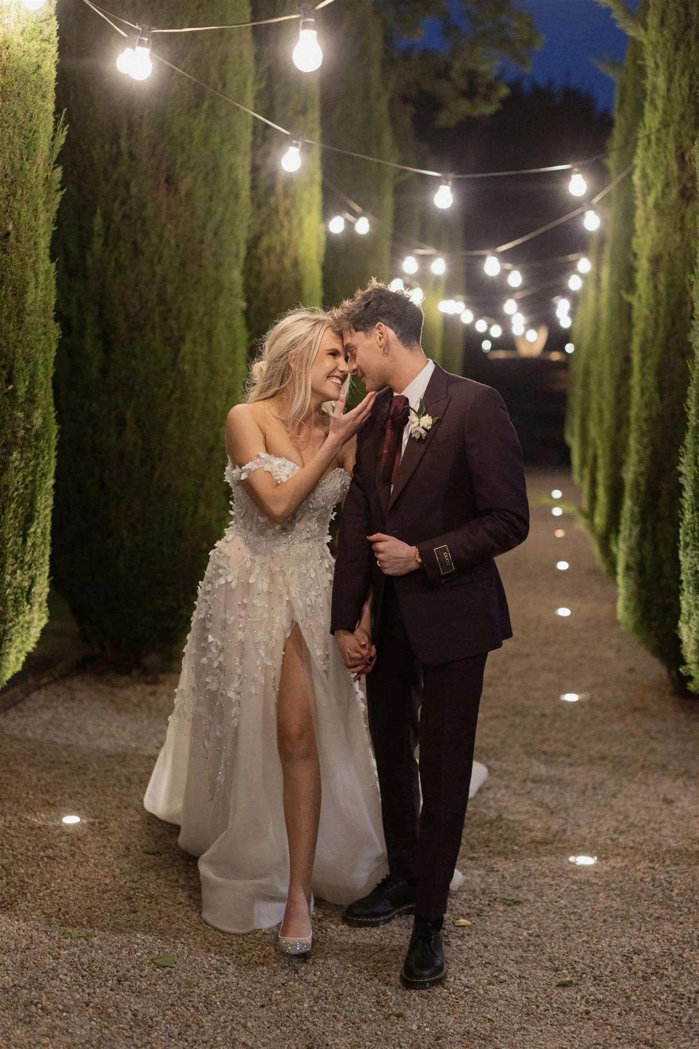 A newlywed couple couples embrace and smile affectionately in an outdoor setting with string lights overhead, surrounded by tall green hedges at night.
