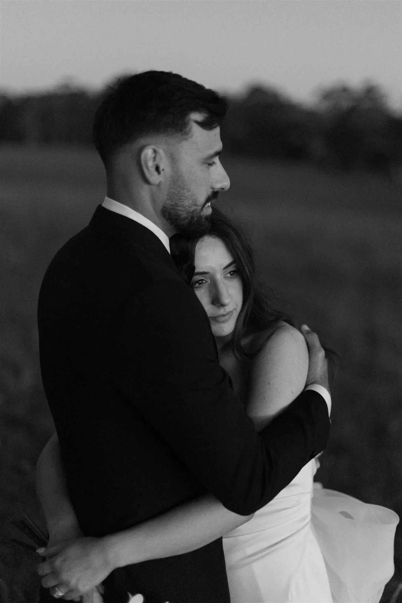 A black-and-white photo of a man and woman embracing outdoors during sunset. The man, dressed in a suit, is holding the woman, who is wearing a strapless dress. They look pensively into the distance.