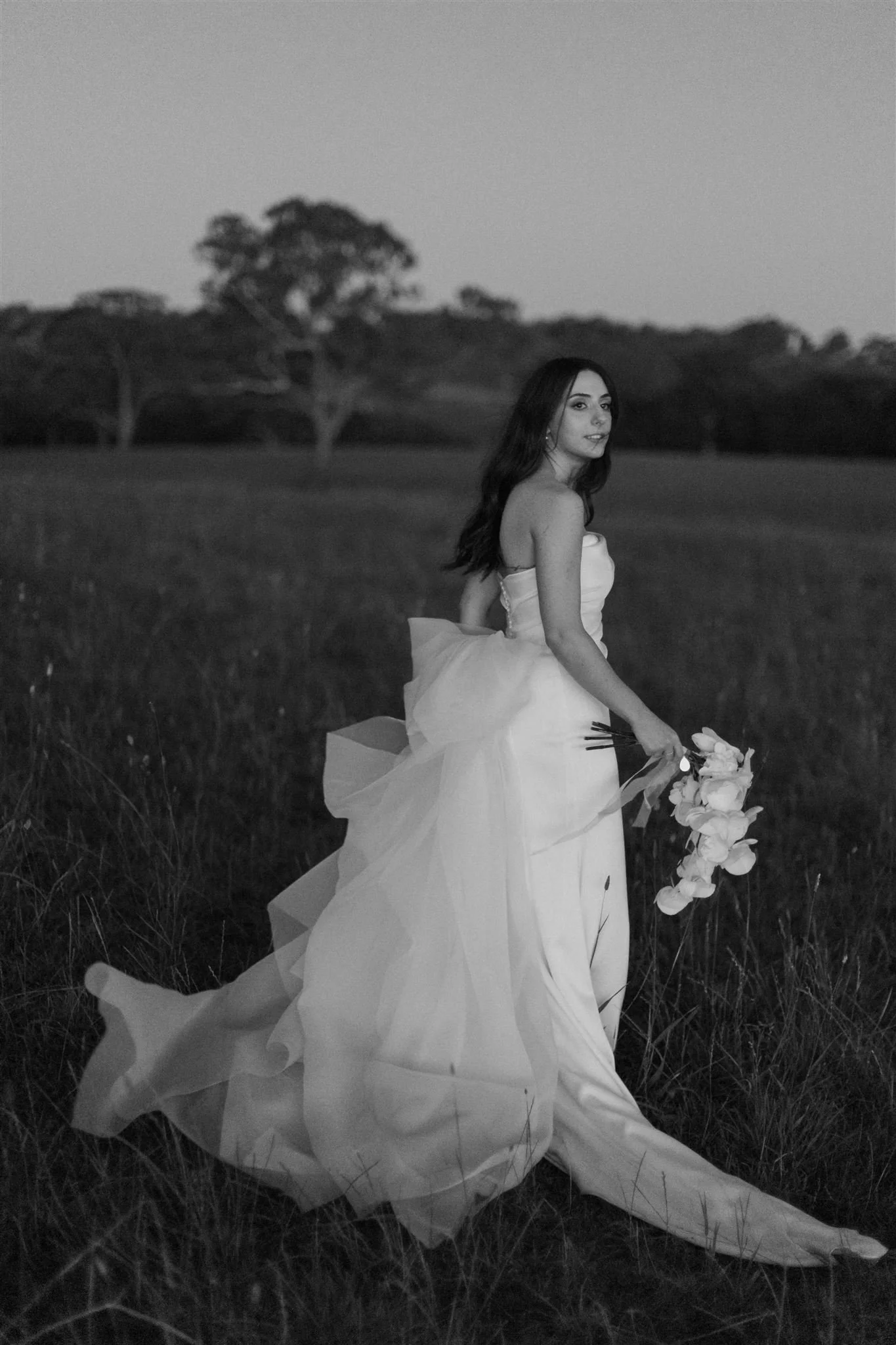 A woman in a wedding dress holding a bouquet stands in a grassy field with trees in the background, captured in black and white. Great Kate made this incredible wedding dress and captured in the fields at Lot100 in the Adelaide Hills by Luke John