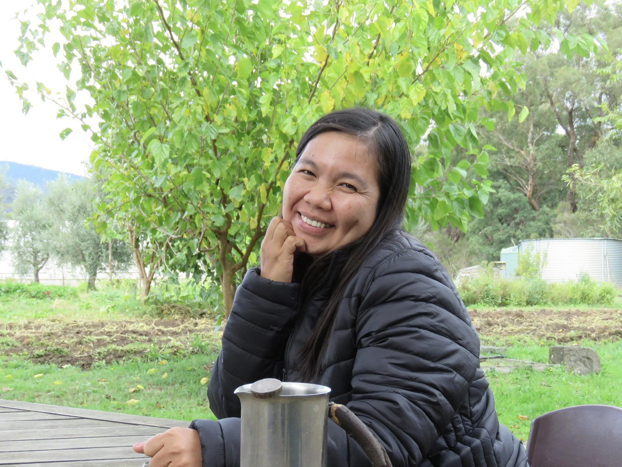 A woman with long dark hair smiling and resting her chin on her hand, sitting at a table outdoors with a lush green background of trees and grass.