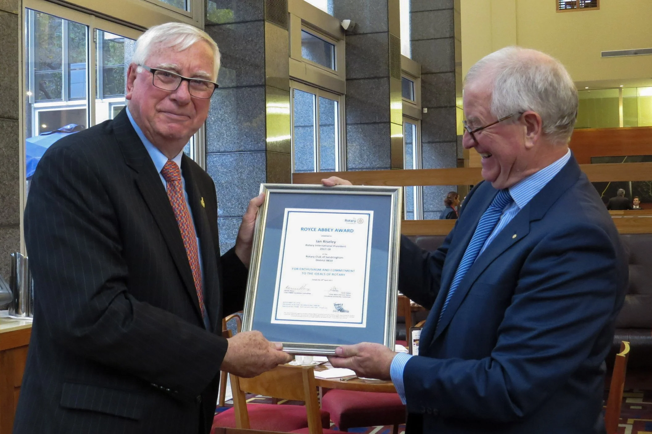 Two elderly men in suits exchanging a framed award, smiling in a formal setting with large windows and wooden furniture.