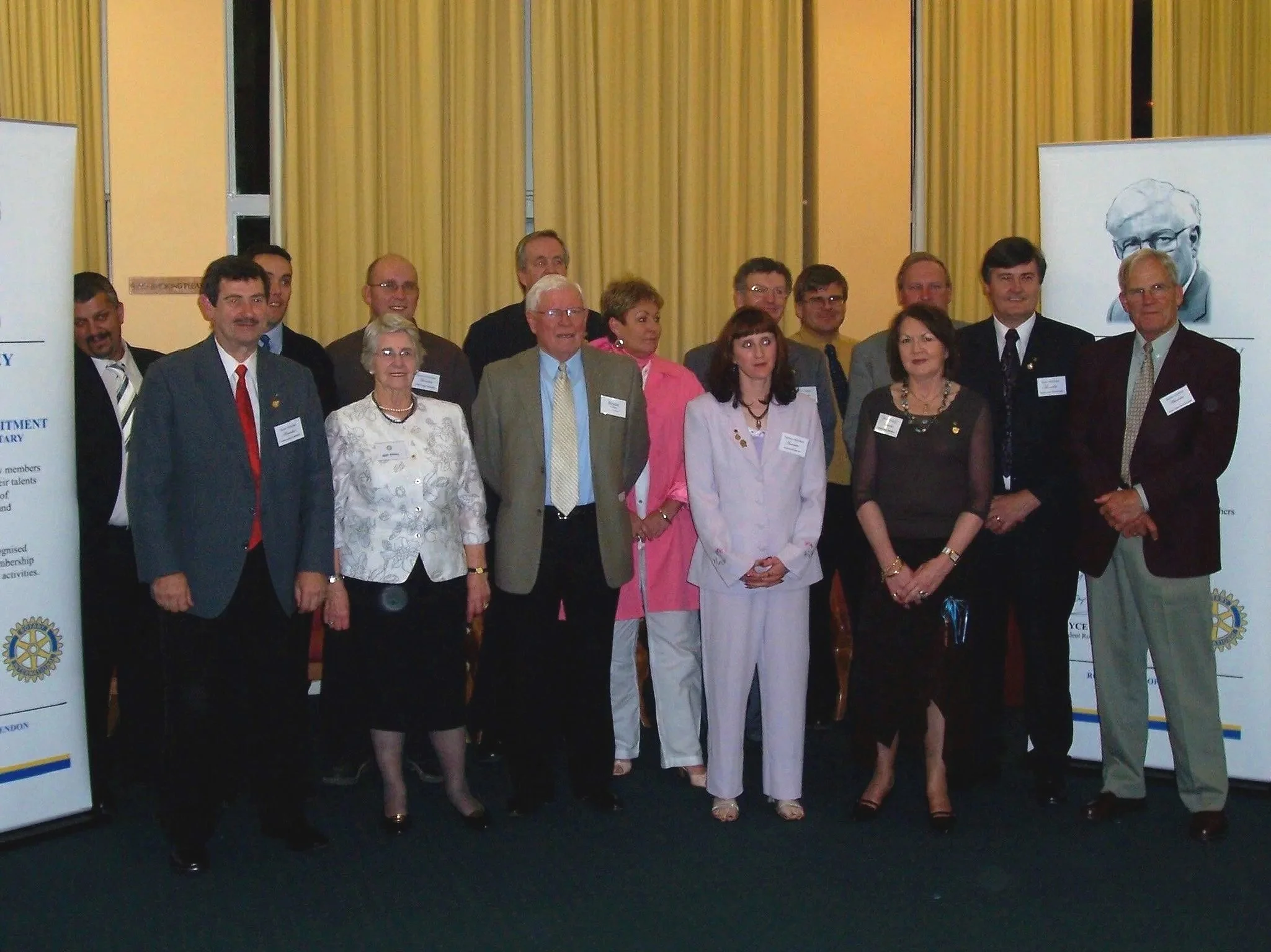 A group of diverse men and women in formal attire posing for a photo indoors, with banners and yellow curtains in the background.