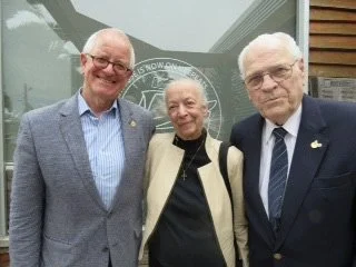 Three individuals standing together outdoors, smiling, in front of a building with a glass window.
