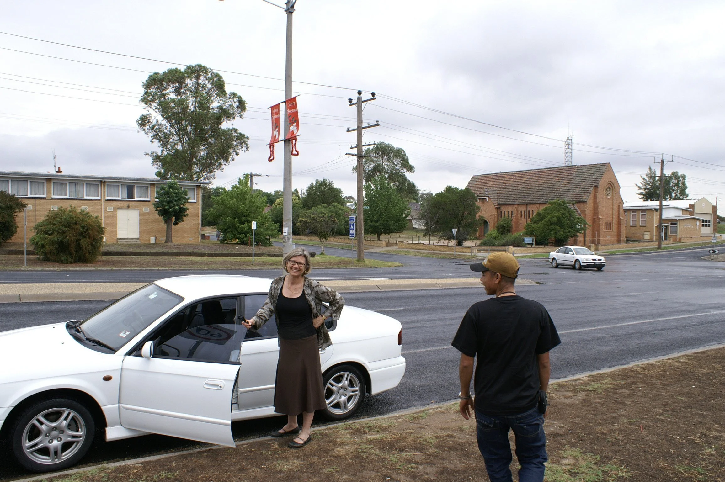A woman with gray hair, glasses, and a black and white patterned jacket standing outside a white car, smiling and holding a car key, while a man in a black t-shirt, dark jeans, and a brown cap stands nearby, on a street with overcast weather and a ch