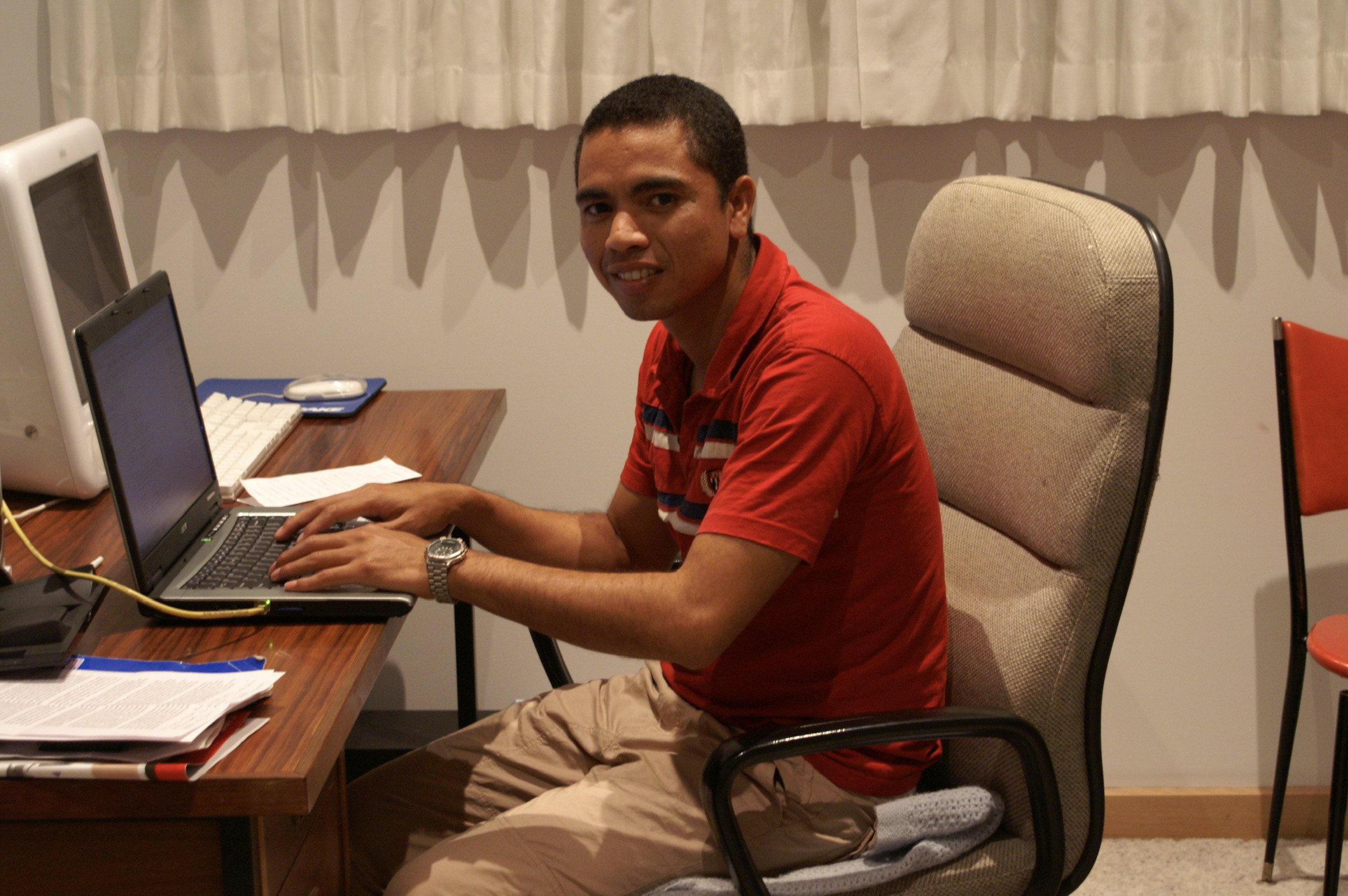 A young man in a red shirt and khaki pants working on a laptop at a desk in a room with beige curtains and a beige office chair.