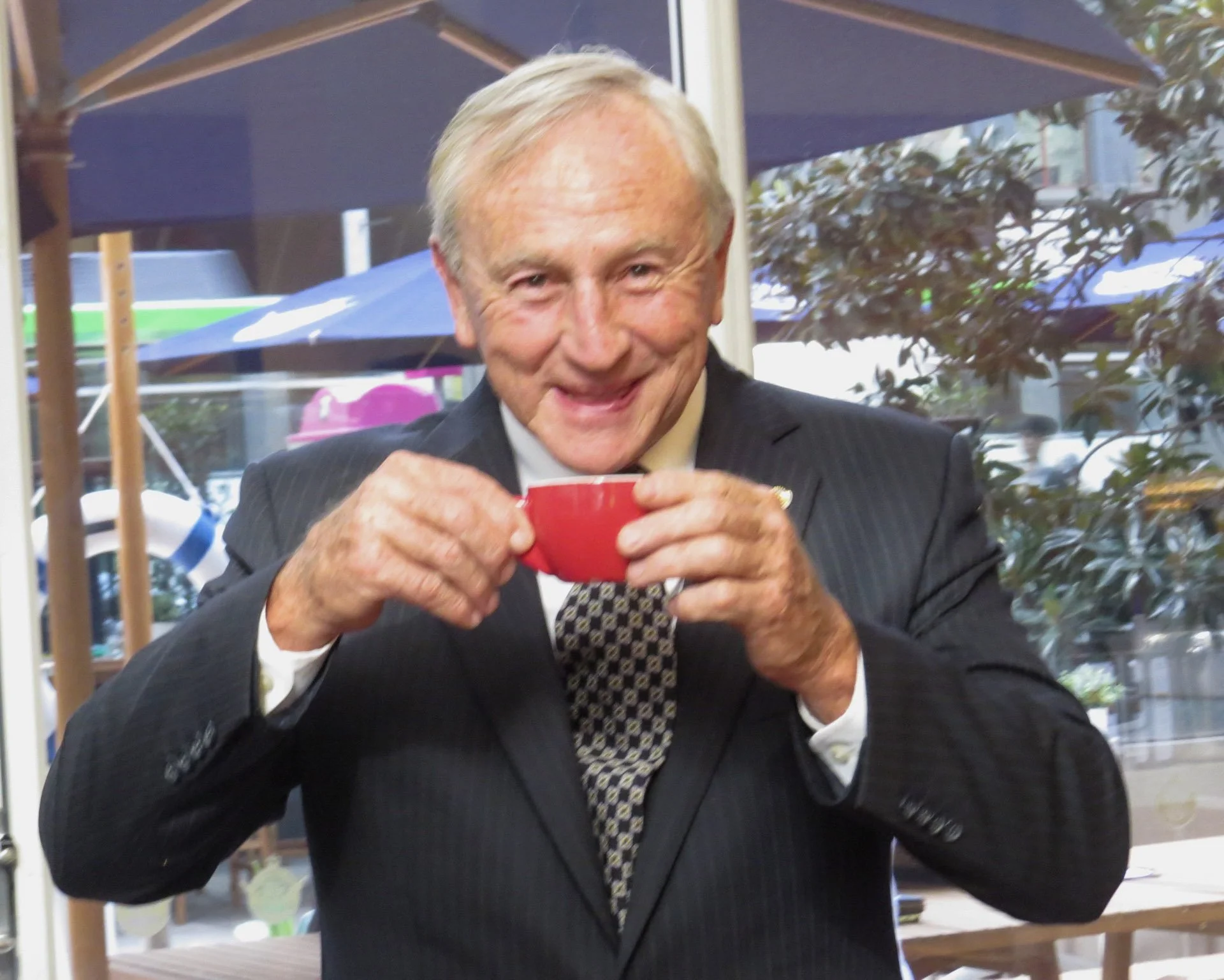 An elderly man in a suit holding a red cup, smiling and enjoying a moment at a cafe.