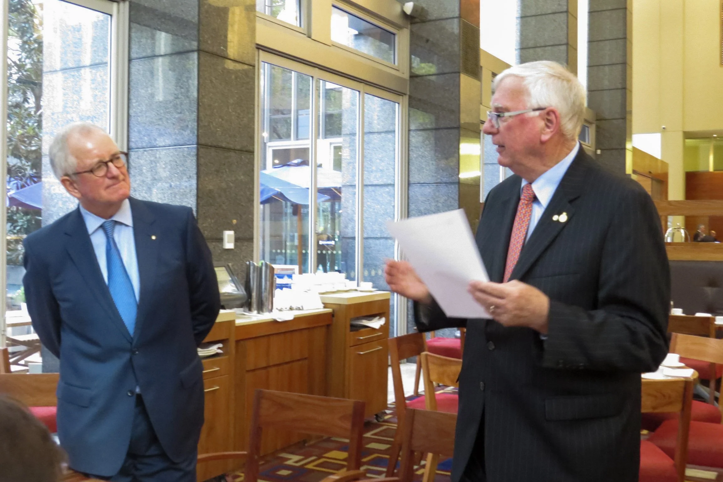 Two older men in suits and glasses are engaged in a conversation in a restaurant or conference room. One man is holding a paper and appears to be speaking, while the other listens. The room has large windows, wooden furniture, and a modern interior d