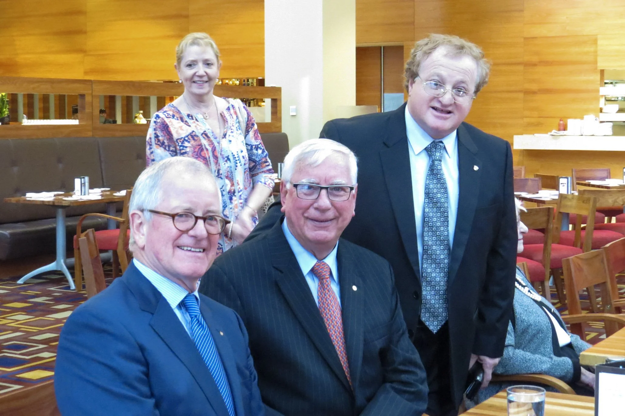 Four people, three men and one woman, are gathered in a restaurant or cafe. The three men are seated at a table, dressed in suits and ties, while the woman stands behind them, wearing a colorful blouse. The background features wooden wall panels and 