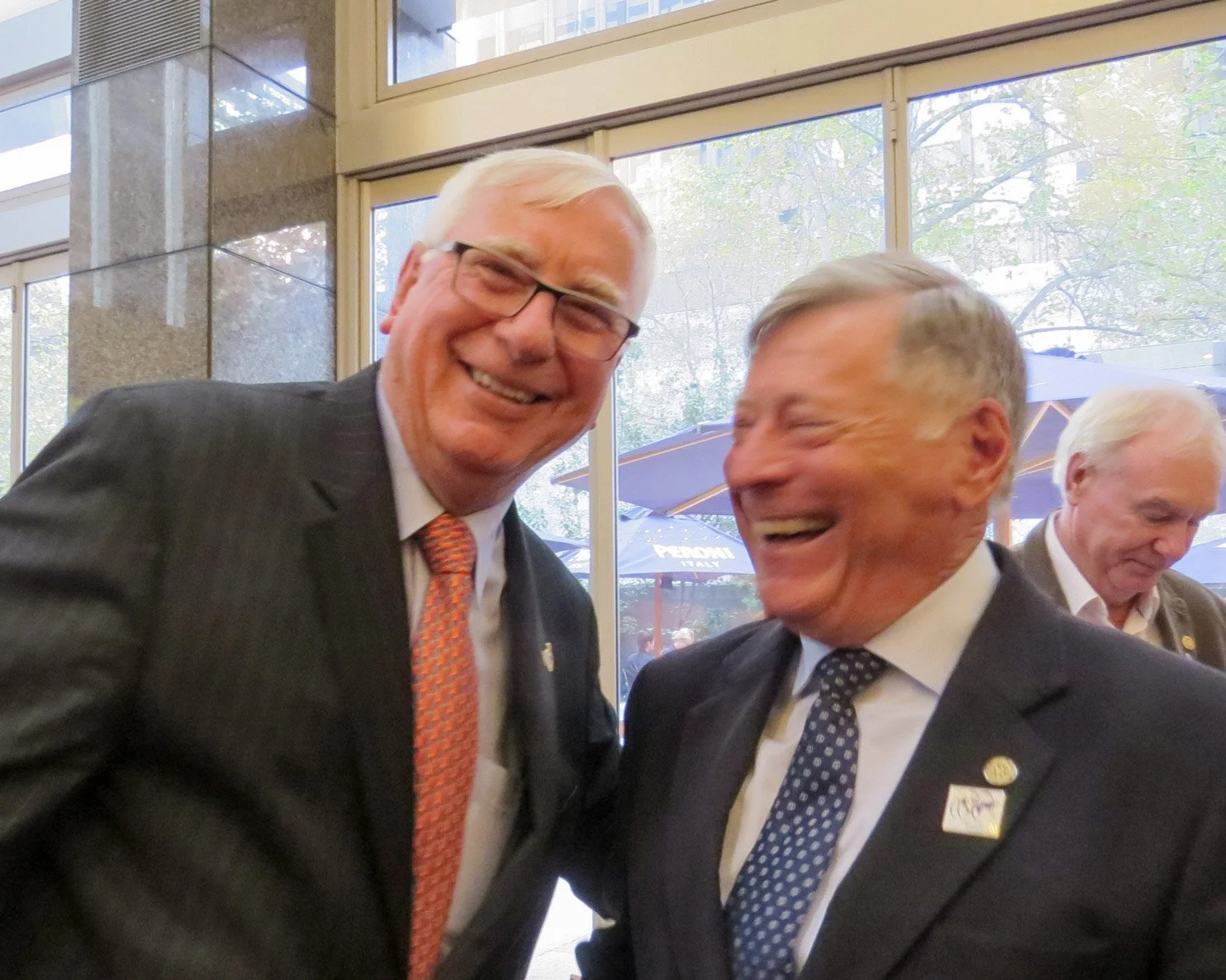 Two men in suits with ties, smiling and engaging in a friendly conversation indoors with a glass window behind them showing trees and umbrellas outside.