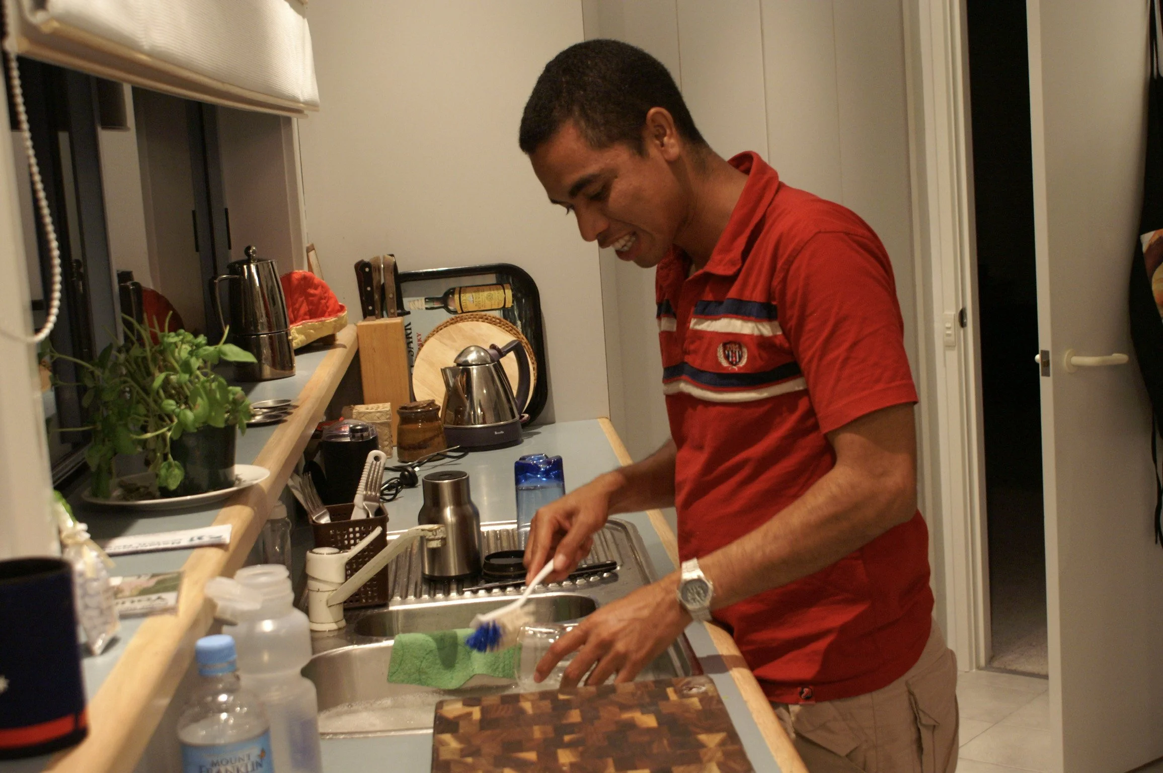 A man in a red shirt washing dishes in a kitchen sink.