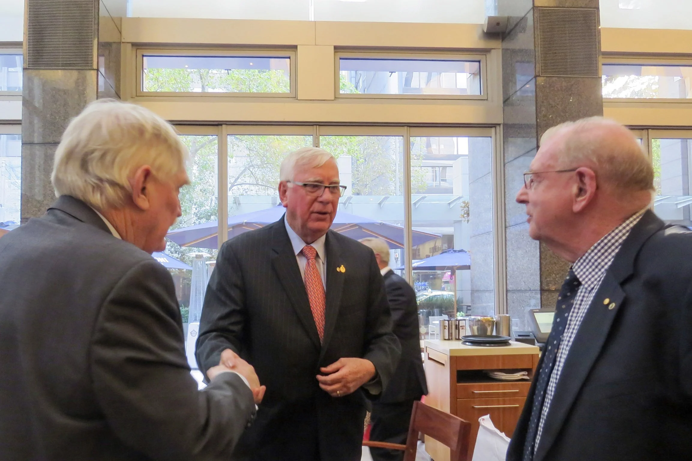 Three men in formal attire are engaged in a conversation indoors near a large window. One man is shaking hands with the other, while the third man stands on the right facing them. The background shows trees outside and a few buildings.