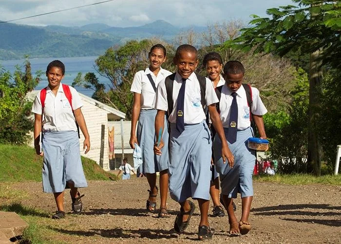 Group of five smiling schoolgirls in uniforms walking outdoors on a dirt path, carrying backpacks and books, with lush green trees, a small building, and a lake in the background.