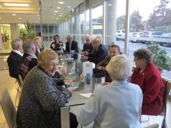 Group of elderly people sitting around a table in a modern, well-lit room with large windows, engaged in conversation and socializing.
