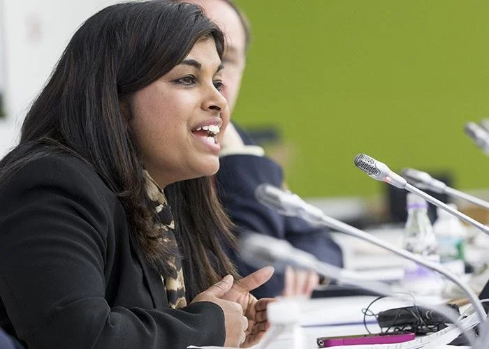 A woman speaking at a panel discussion or meeting with microphones in front of her