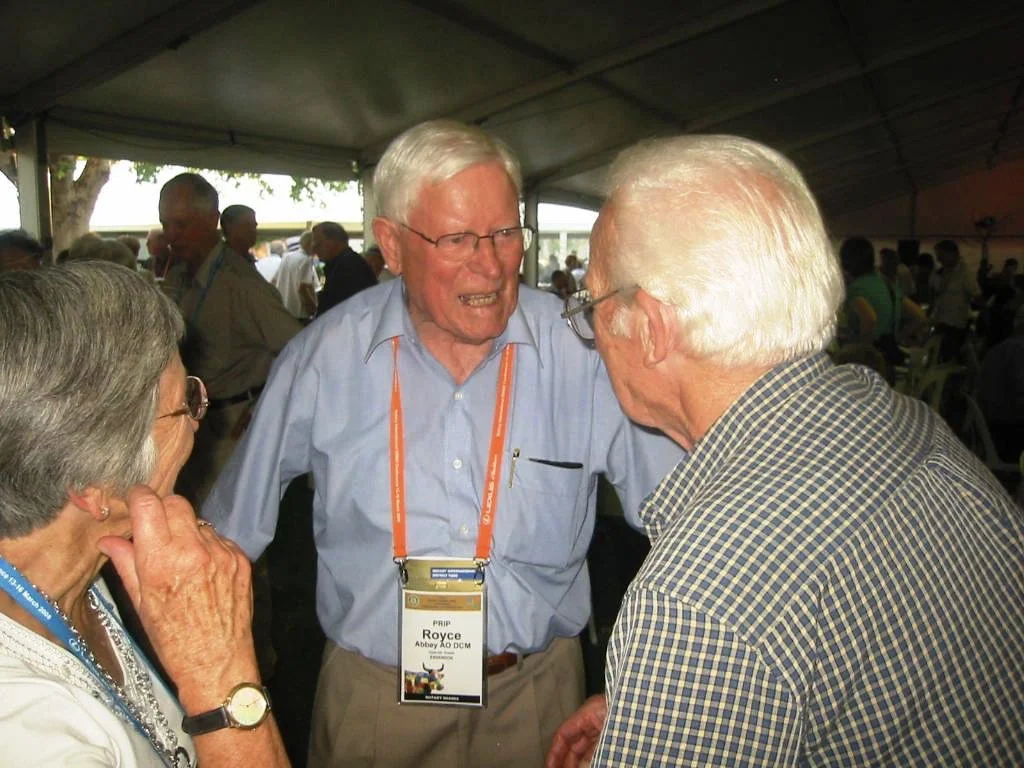 Three elderly individuals engaged in an animated conversation under a large tent at a conference or event.