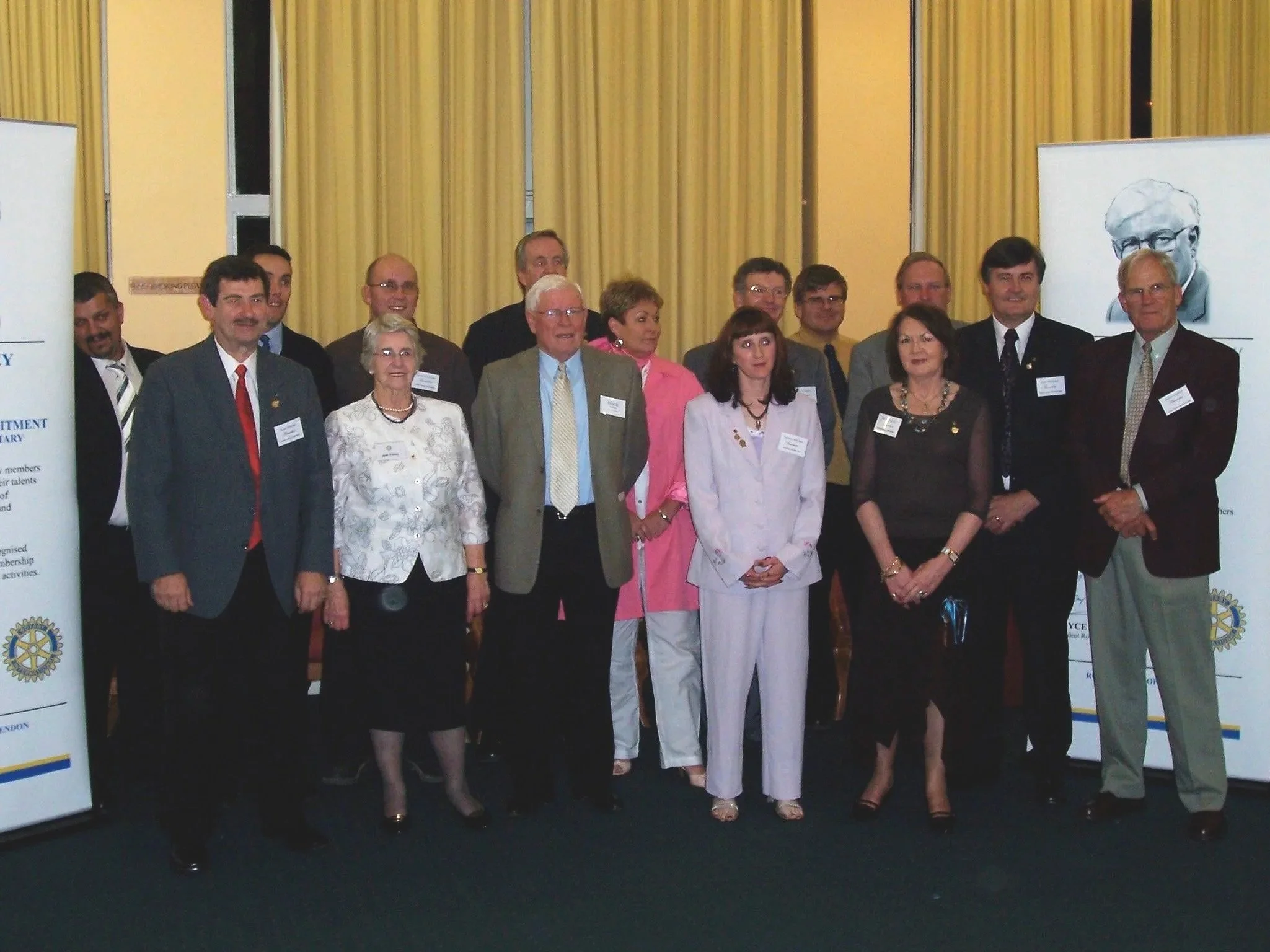 Group of professionals at a conference or formal event, standing in front of yellow curtains and banners with Rotary International logos.