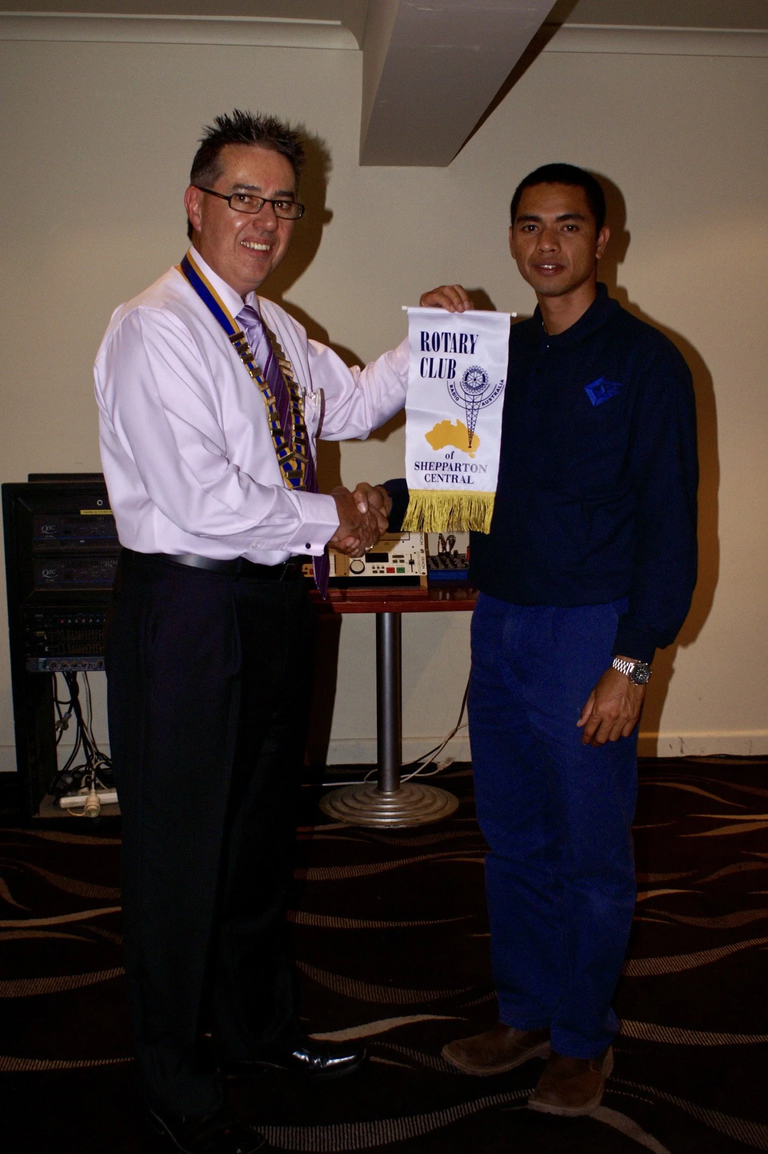 Two men shaking hands at a Rotary Club event, one holding a Rotary Club flag that says 'Rotary Club of Shepparton Central'.