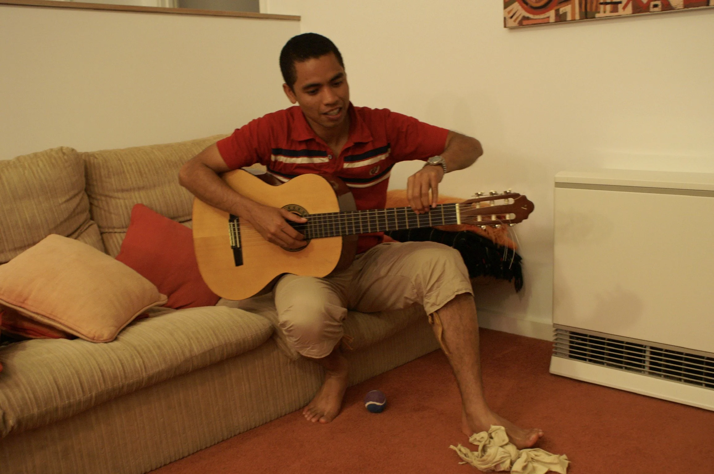 A young man with short dark hair, wearing a red striped polo shirt and beige pants, sitting on a beige couch, playing an acoustic guitar. There are pillows on the couch, and a crumpled cloth on the carpet near his foot. The background shows a plain w