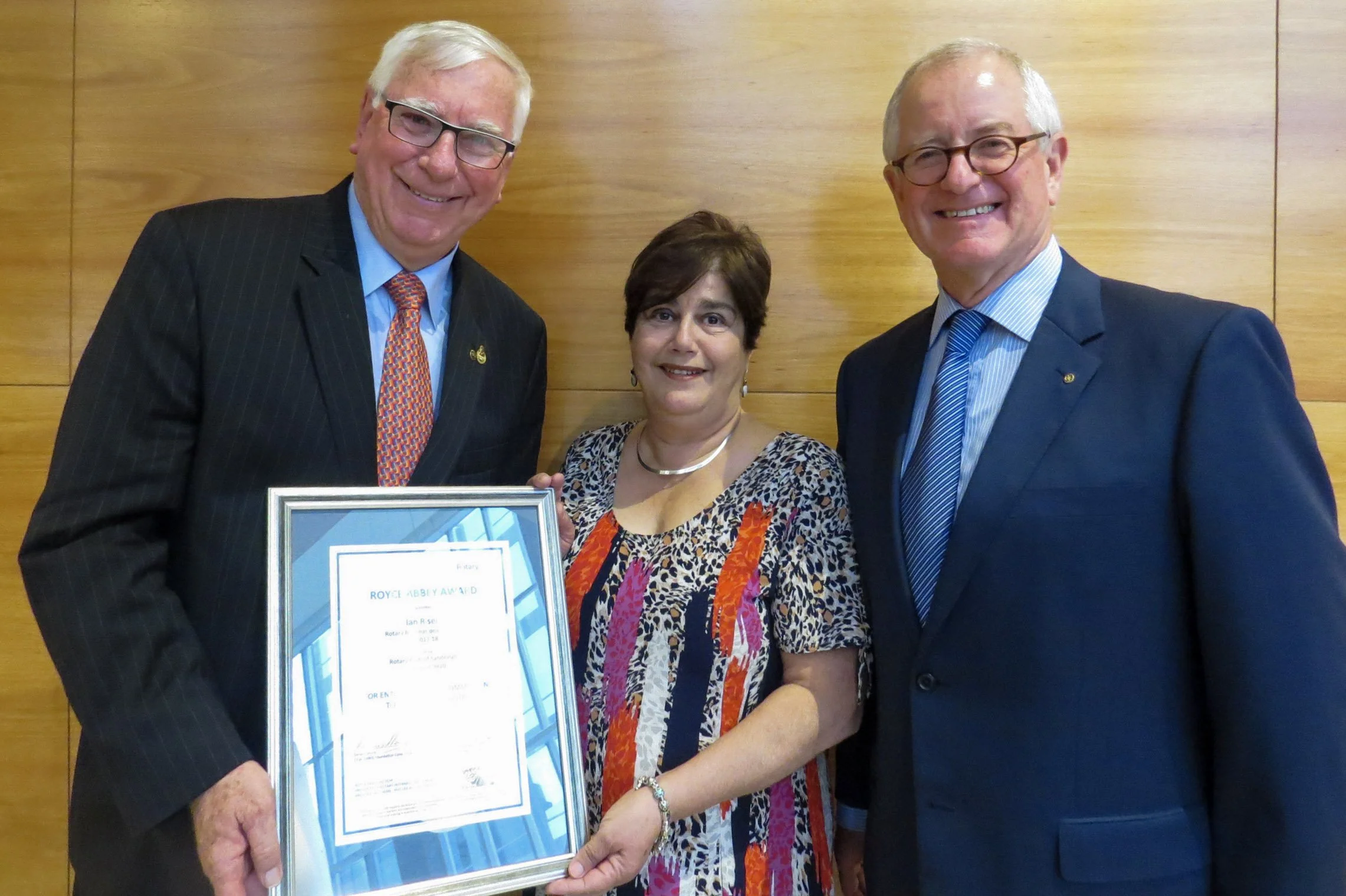 Three people smiling at the camera, two men in suits and a woman in a colorful dress, holding a framed certificate with a wooden wall in the background.