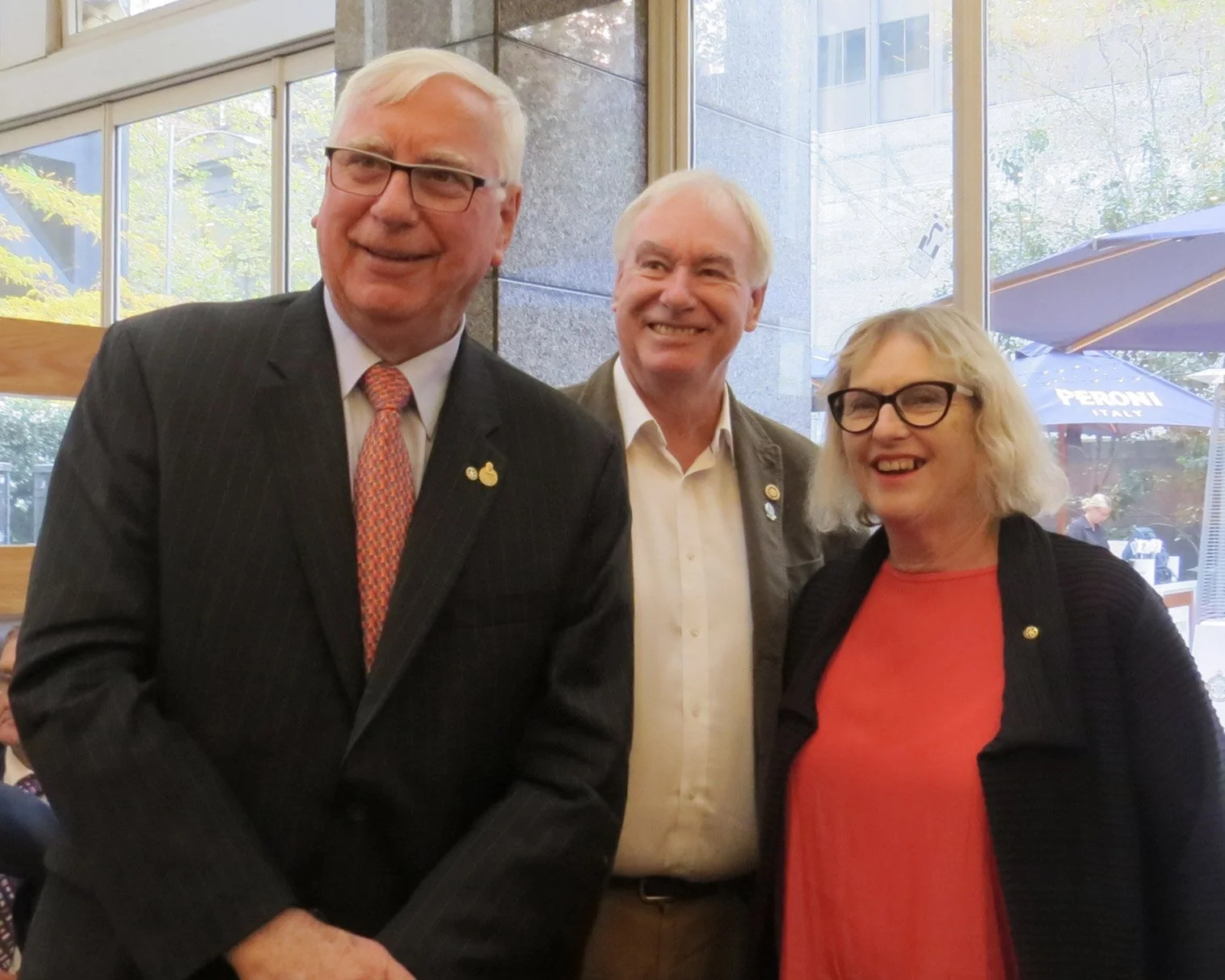 Three people standing inside near large windows, smiling, dressed in business casual attire; two men and one woman.