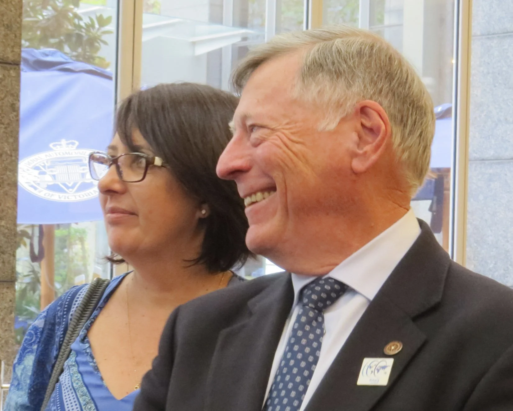 A smiling man in a suit with a pin and a woman wearing glasses and a blue top, sitting indoors during daytime.