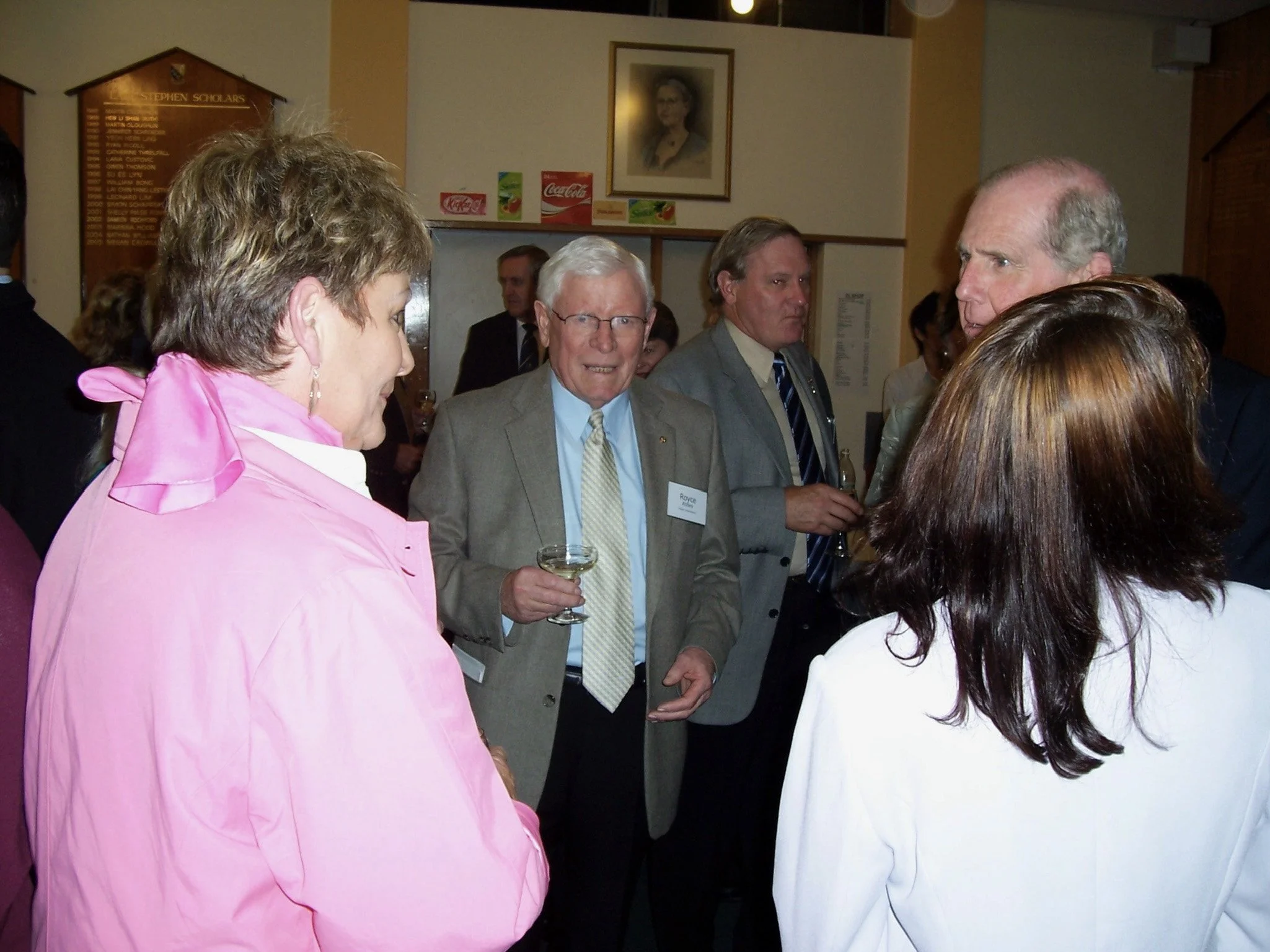 A group of people dressed in formal and semi-formal attire socializing at an indoor event, with a woman in a pink jacket and a woman in a white blazer in the foreground, and four men, one holding a glass of wine, engaged in conversation.