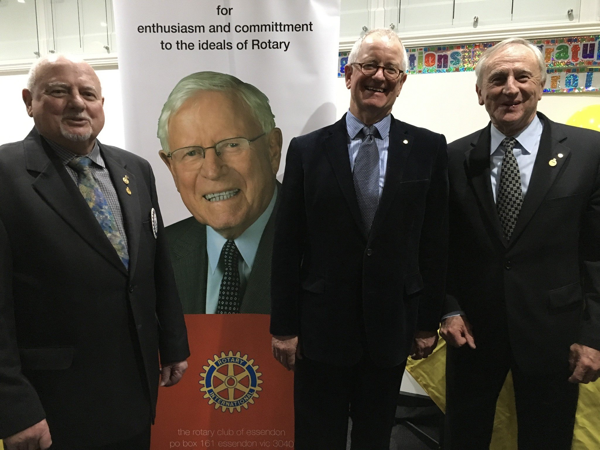 Four men in suits standing together at a Rotary event, with a large poster of a smiling man and the Rotary logo in the background.