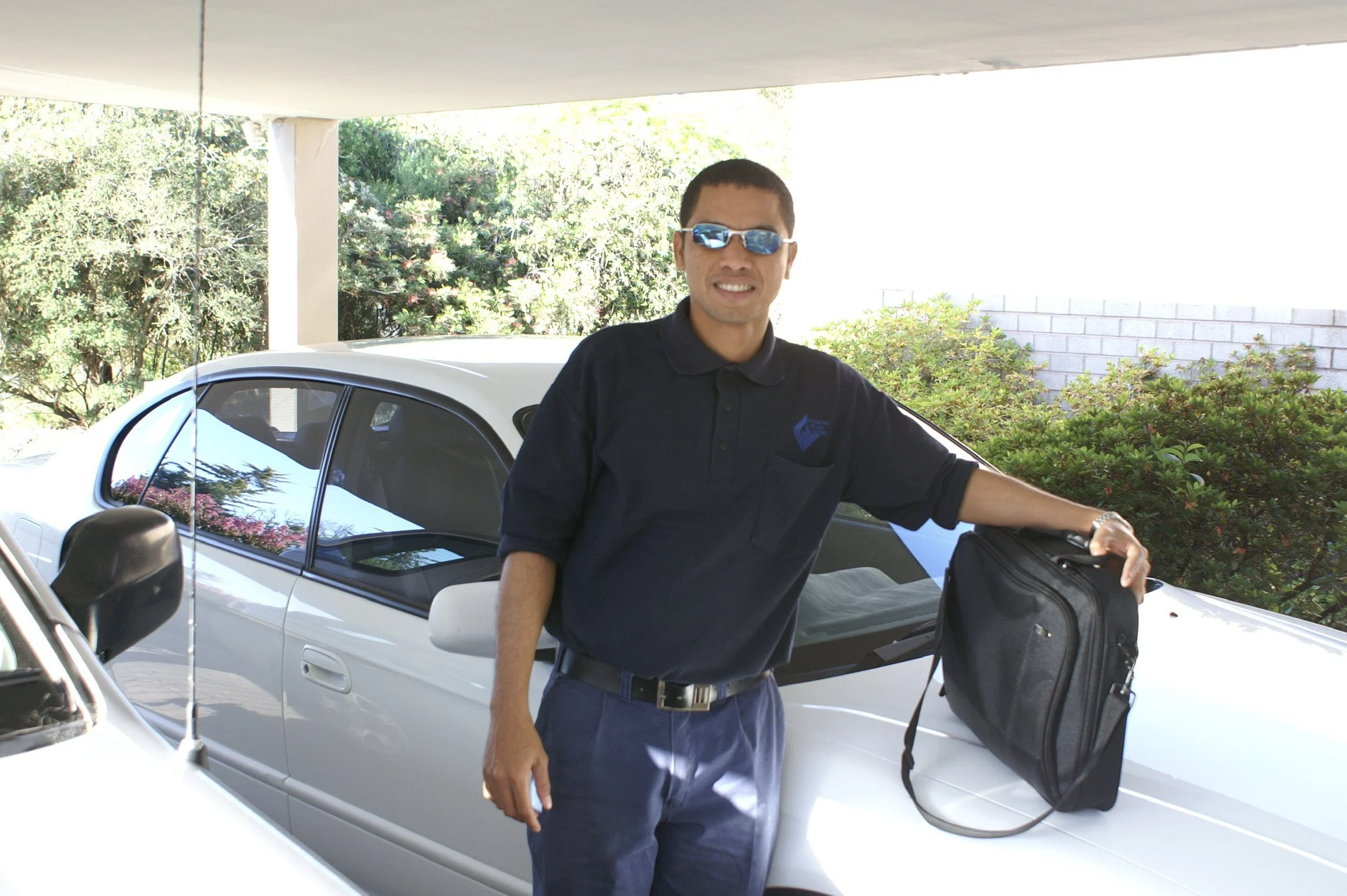 Man in sunglasses and dark polo shirt leaning on a white car with a black bag on the hood.