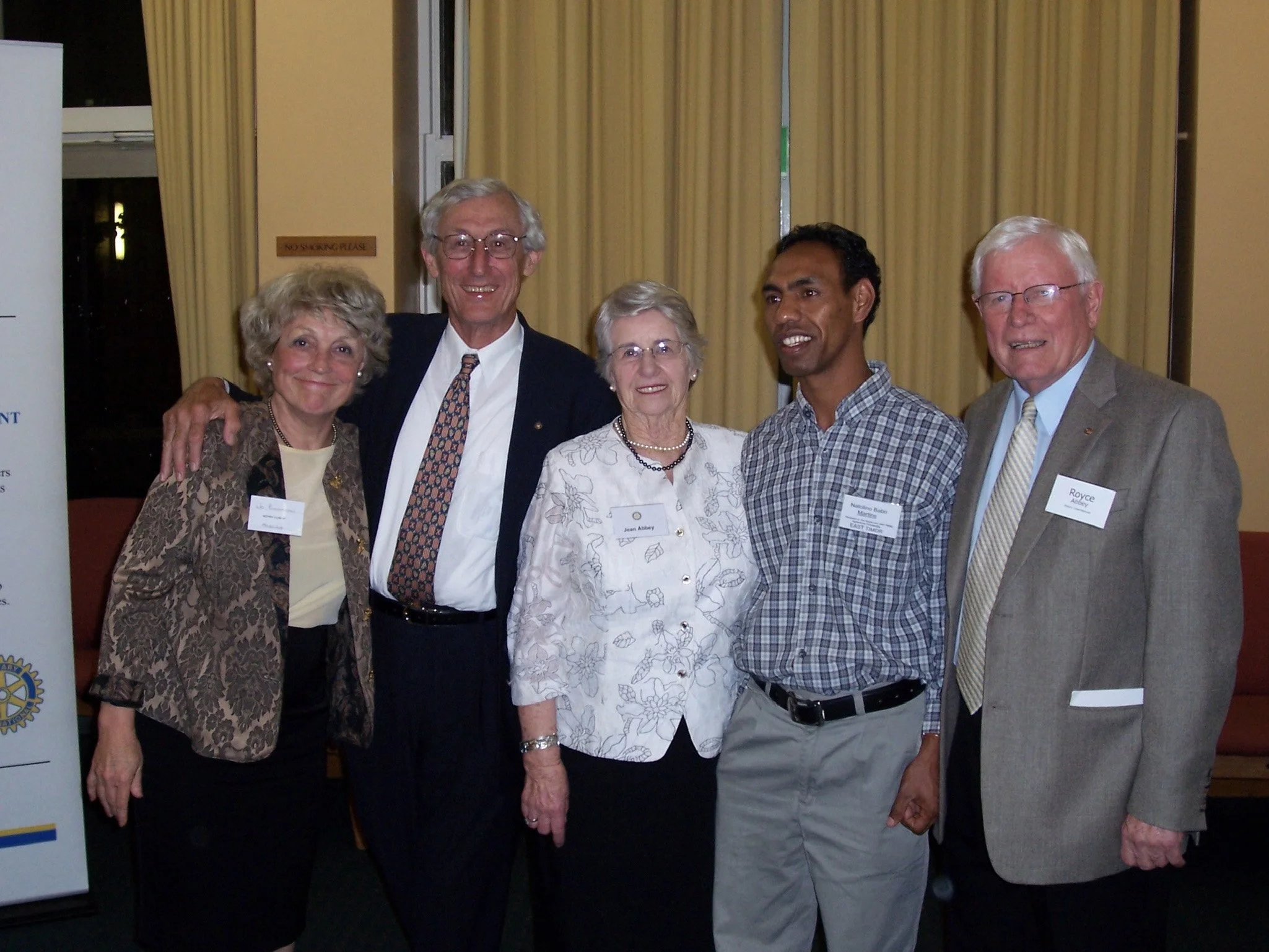 Six people standing together, smiling, at a formal event, with name tags, inside a room with curtains and a banner on the left.