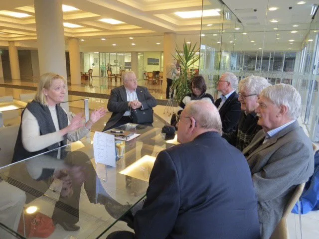 A group of seven adults seated around a glass-topped conference table, engaged in a discussion inside a modern office or meeting space with large windows and indoor plants.