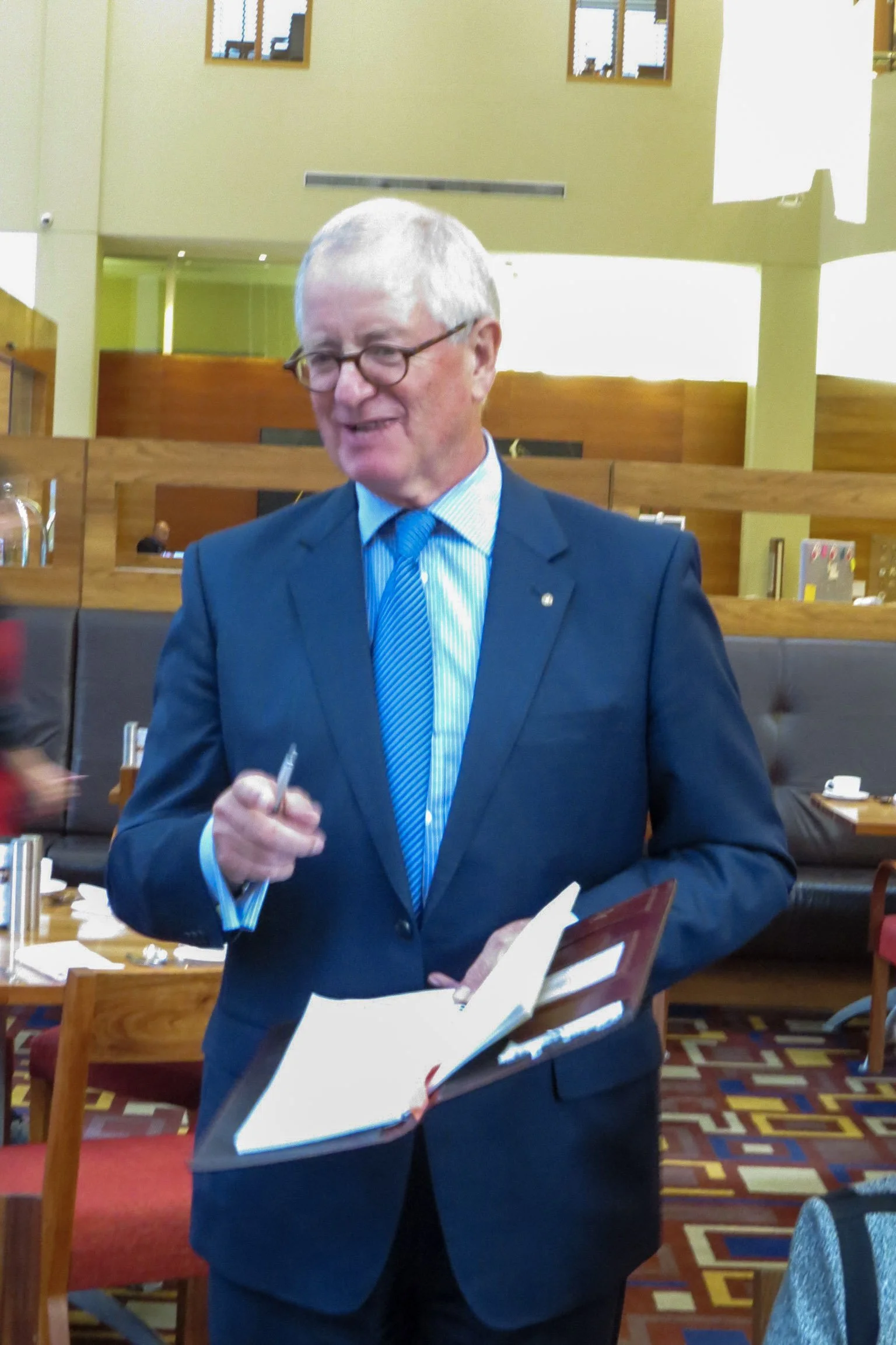 A man in a blue suit and glasses holding a notebook and pen, standing in a restaurant or cafe setting.